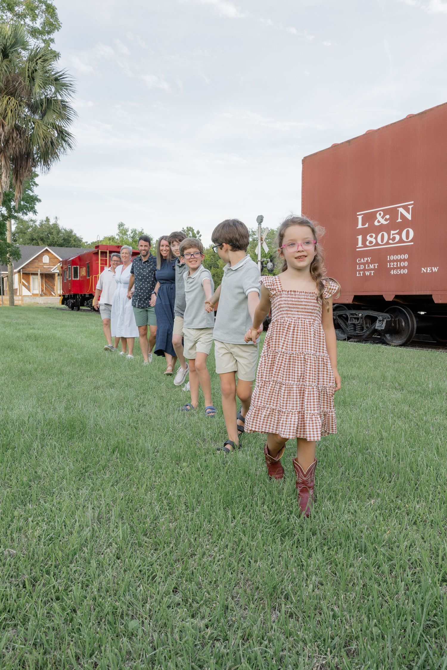Children and adults walk in a line on green grass beside a vintage L&N railroad freight car outdoors.