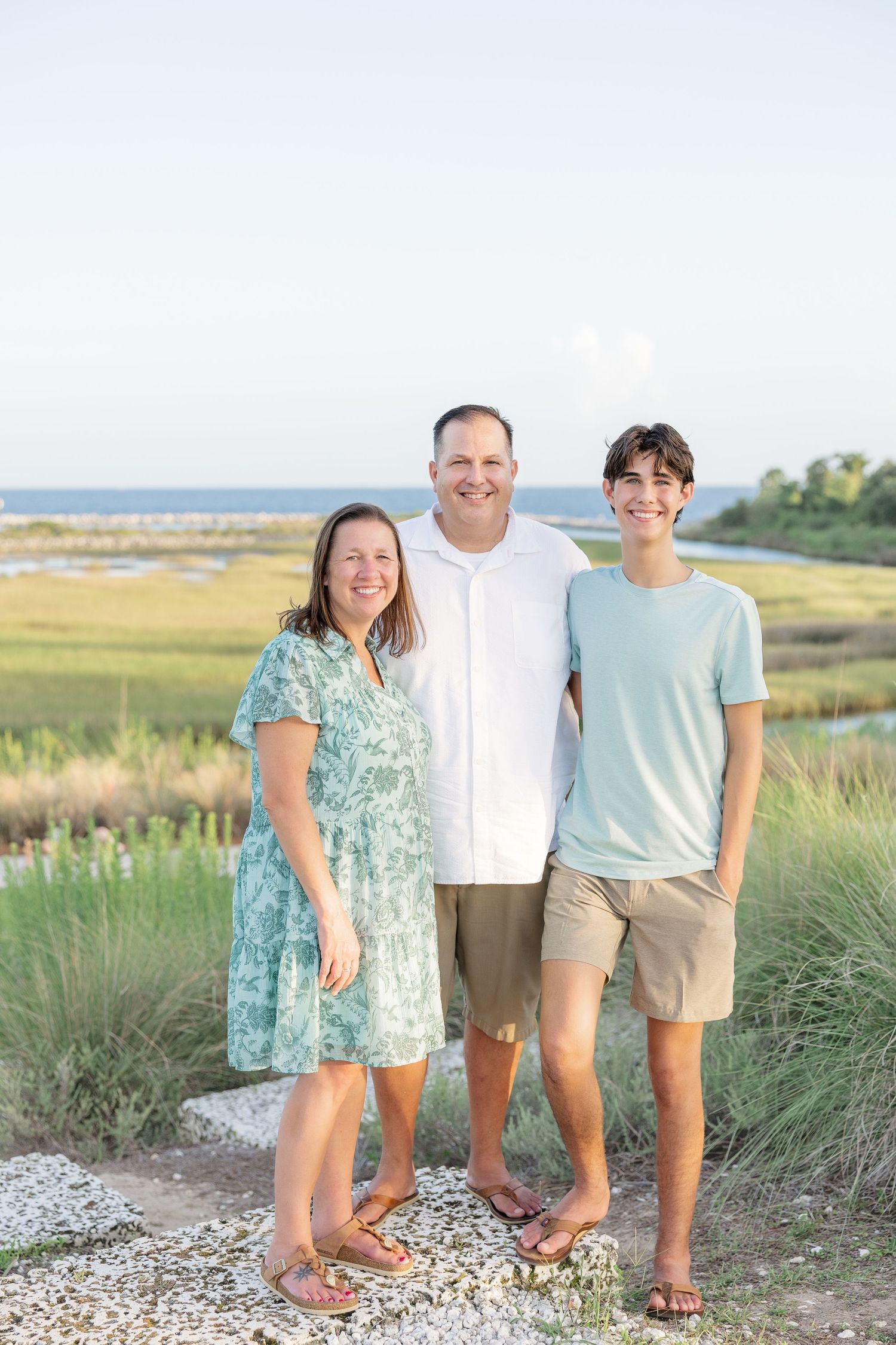 A smiling family of three poses outdoors near a coastal marsh, wearing casual summer attire on a sunny day.