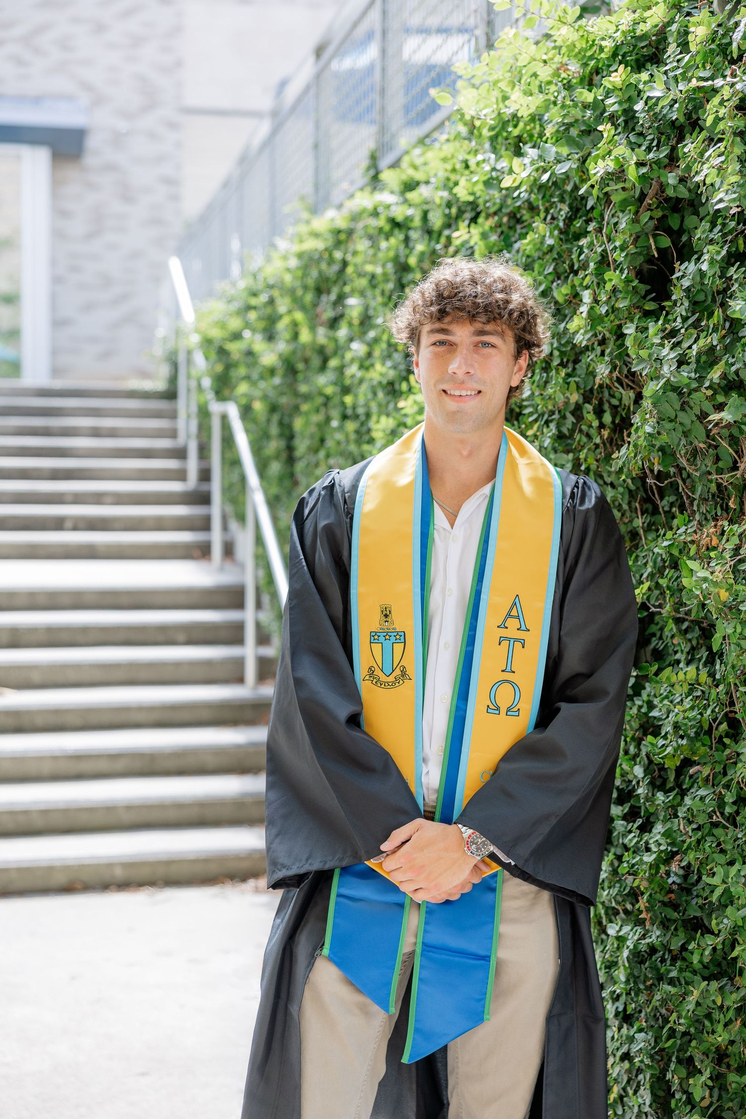 Young man in black graduation gown with gold and blue fraternity stoles smiling outdoors near ivy-covered wall.