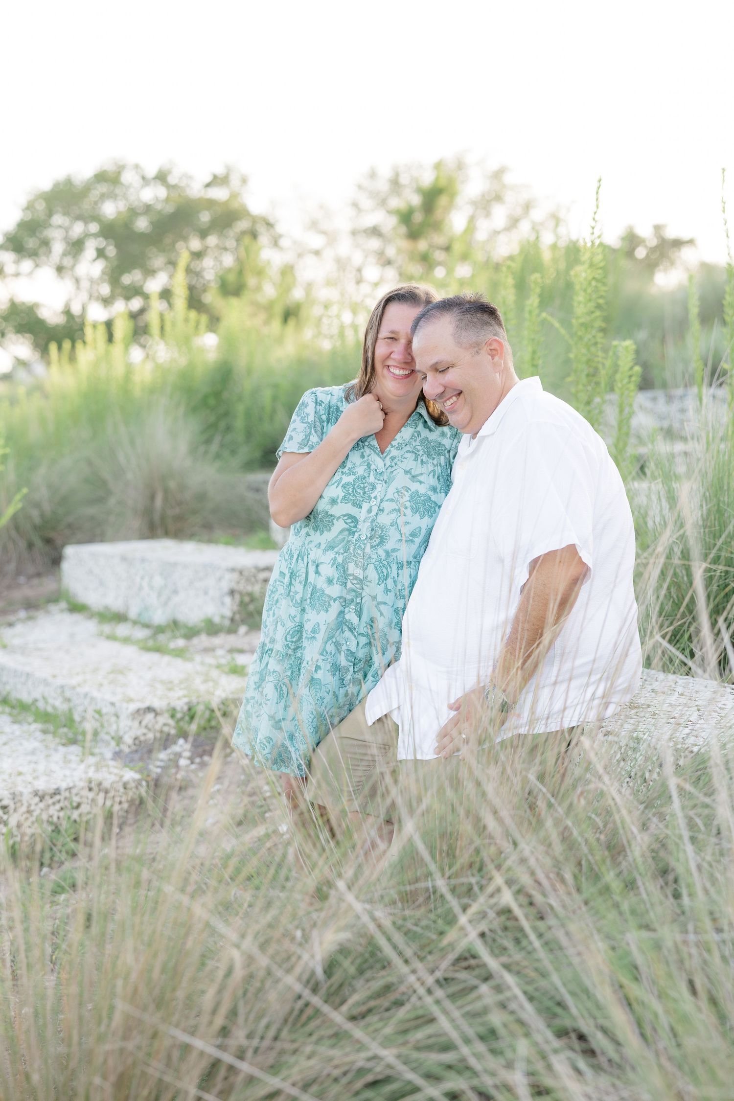 A happy couple laughs together outdoors among tall green grasses, woman in teal dress and man in white shirt.