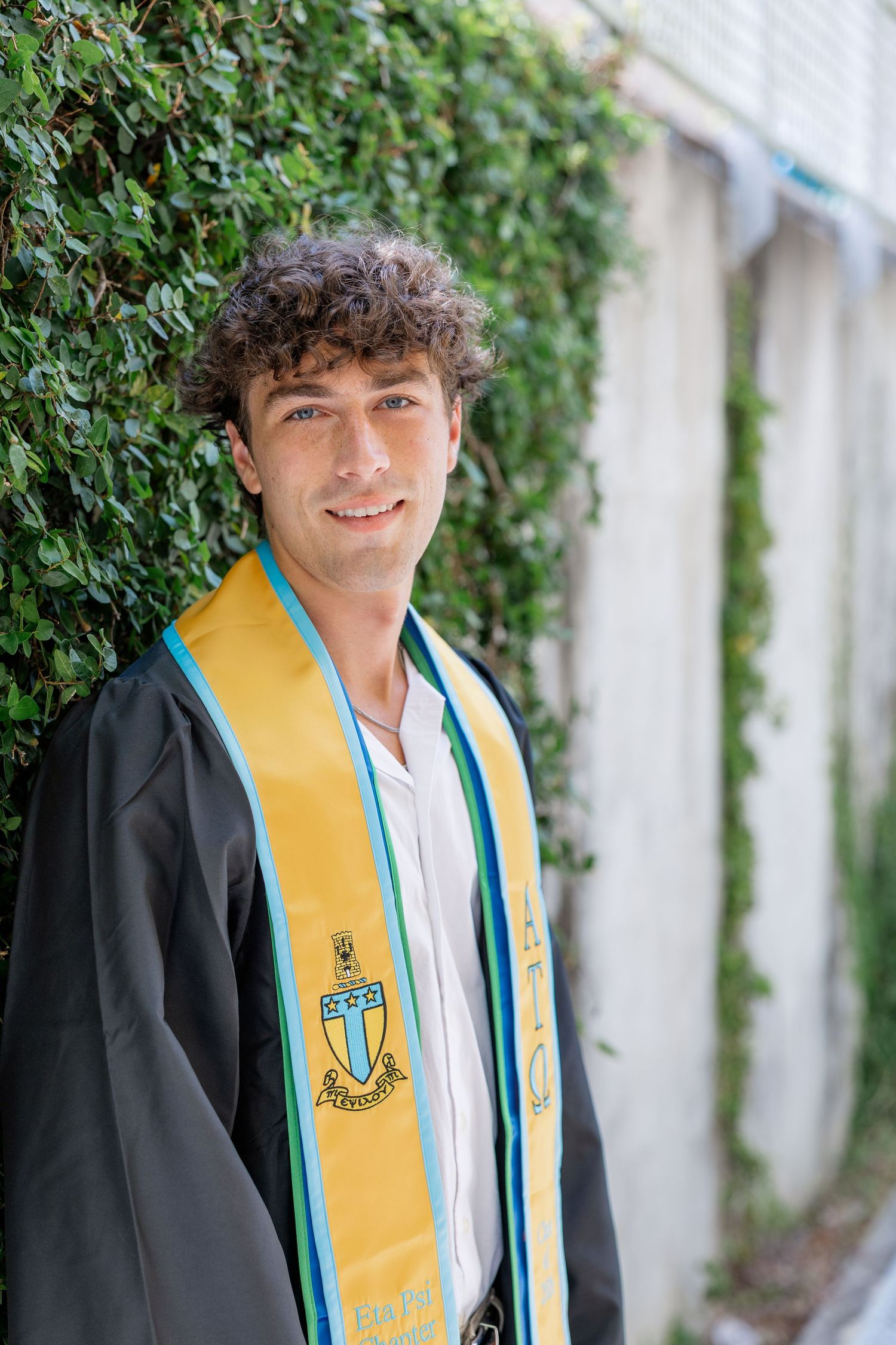 Young male graduate smiling in black gown and gold UWF stole against ivy wall.