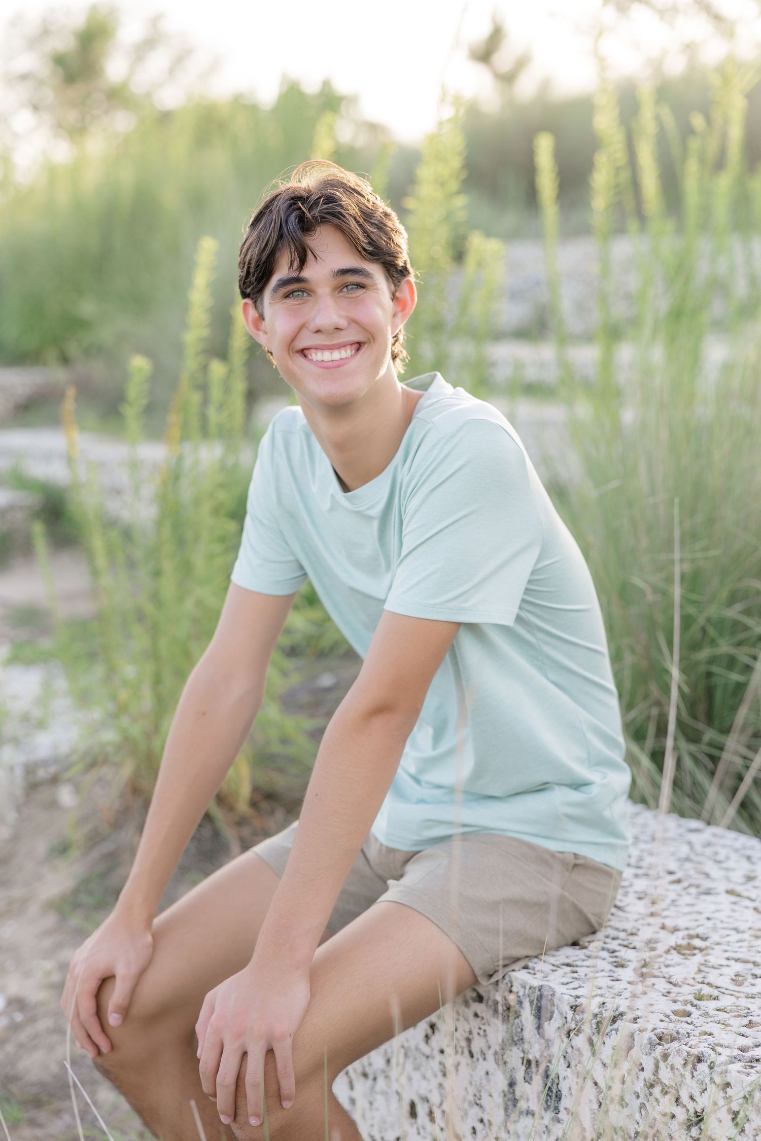 Smiling teenage boy in mint green t-shirt and khaki shorts sitting on stone wall outdoors with green grass background.