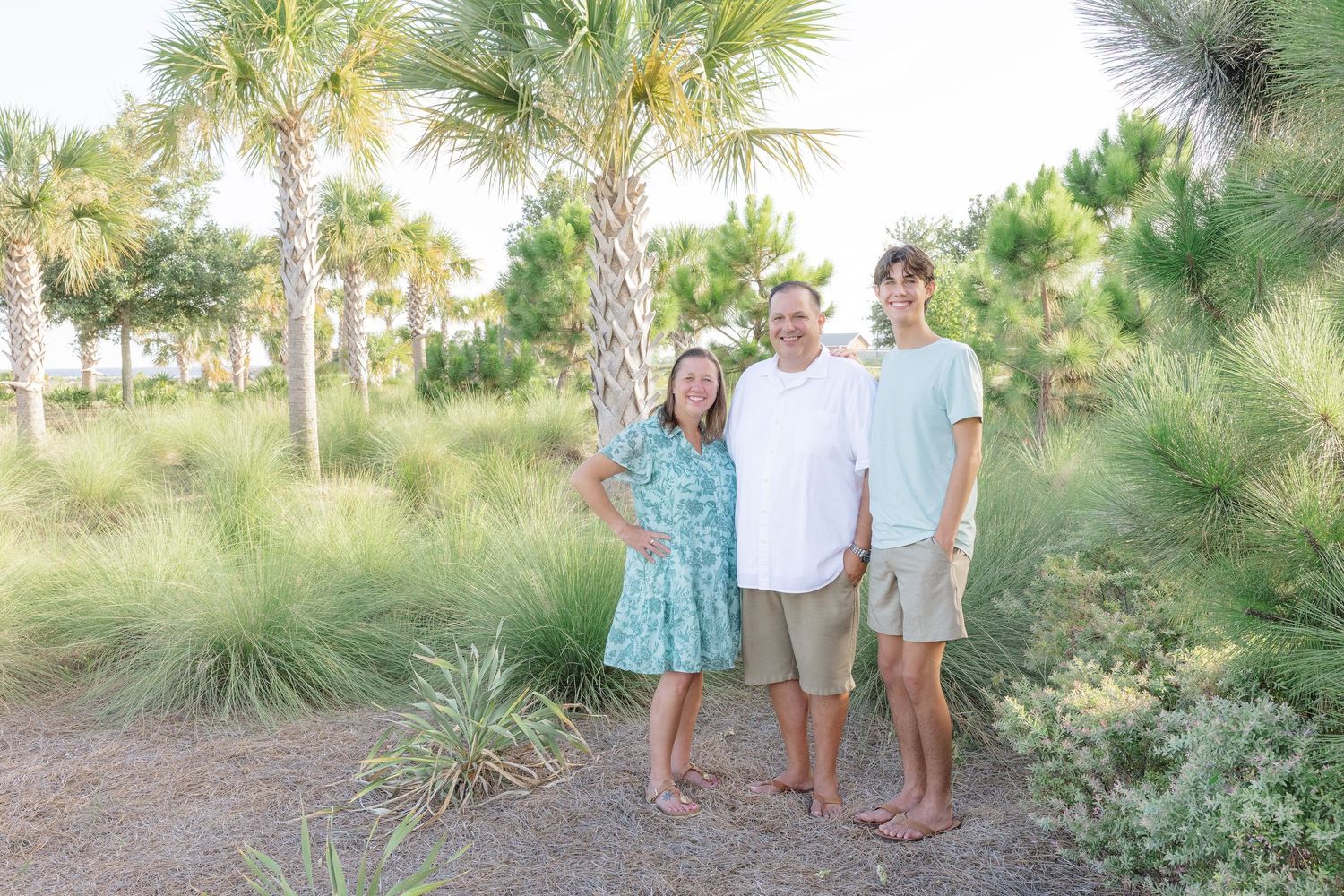 Family of three smiling outdoors among palm trees and lush greenery in a tropical setting.