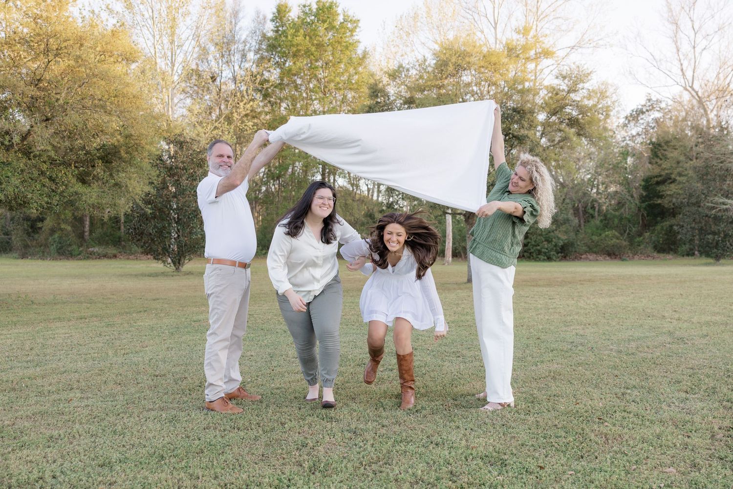 Family of four playing outdoors, holding a white blanket over two girls running underneath in a sunny green park.