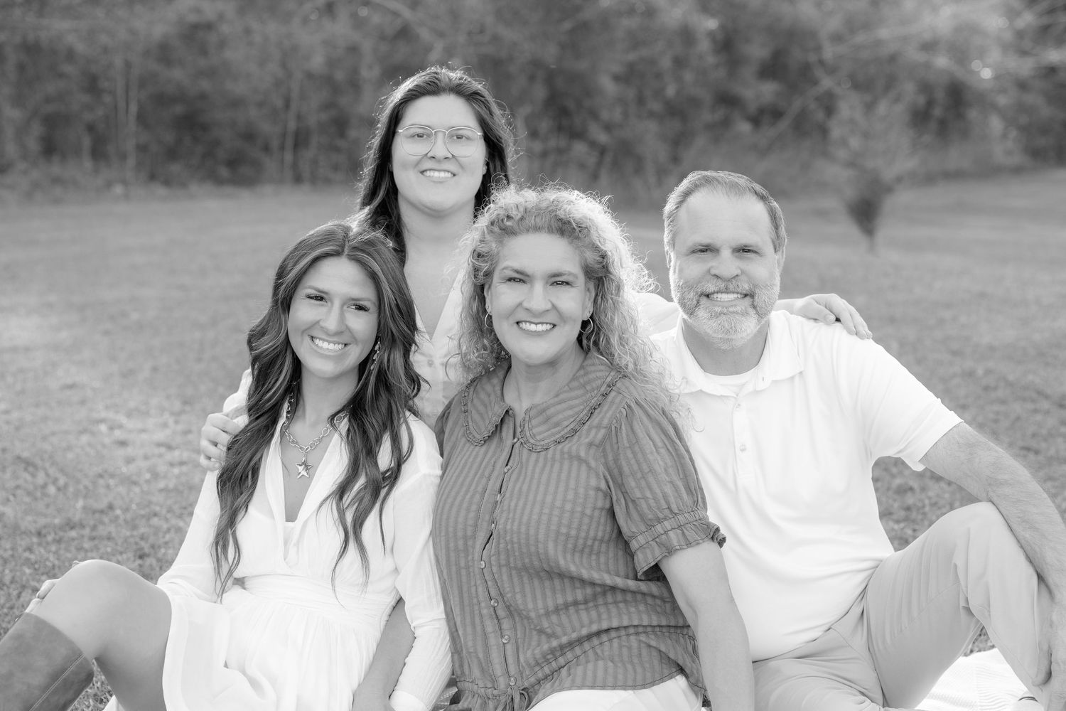 Black and white family portrait of four smiling people sitting outdoors on grass in casual white attire.