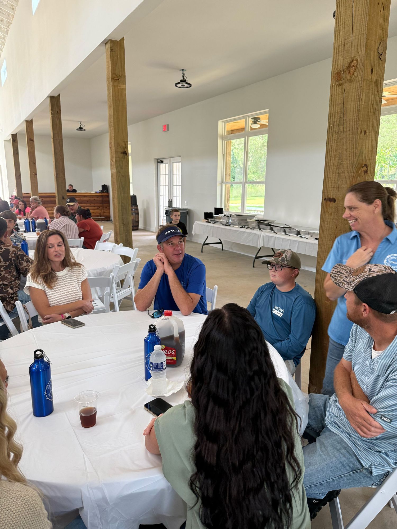 People seated around tables in a bright community hall with wooden posts and white tablecloths.