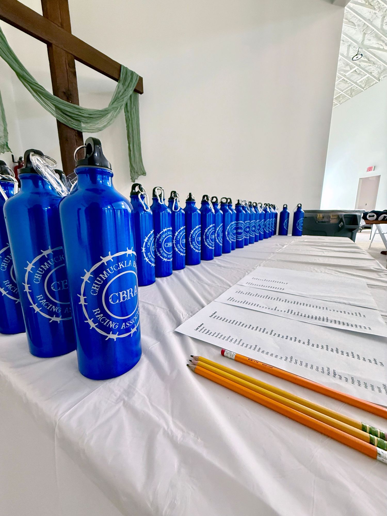 A line of identical blue promotional water bottles and pencils arranged diagonally on a white tablecloth.