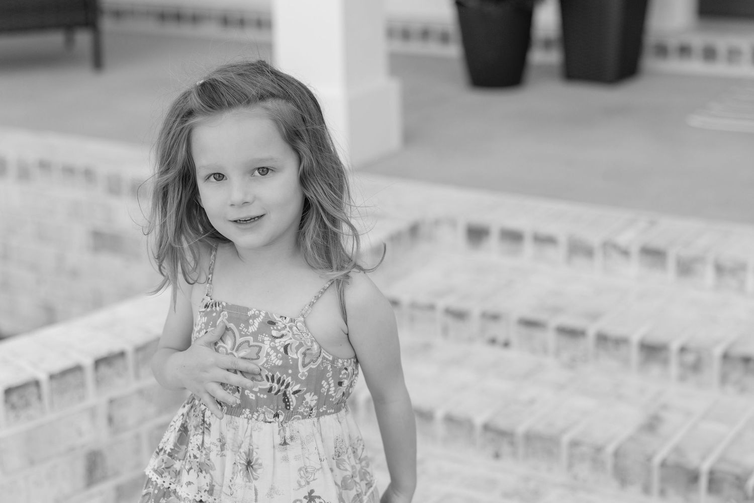 Young girl in floral sundress smiling outdoors near brick steps in black and white photo.