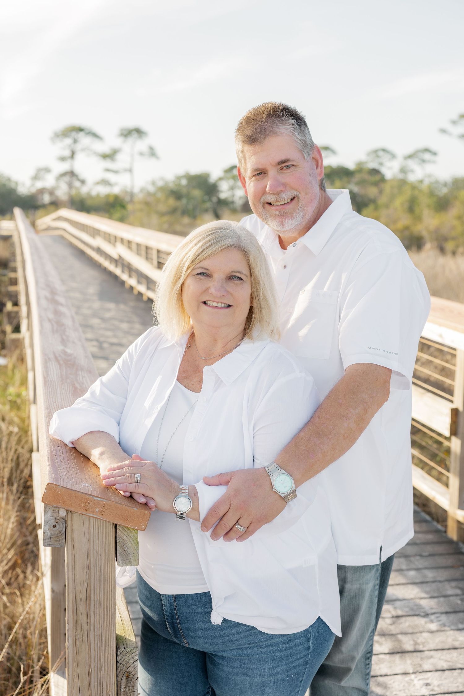 A couple in white shirts stands together on a wooden boardwalk surrounded by coastal vegetation on a sunny day.