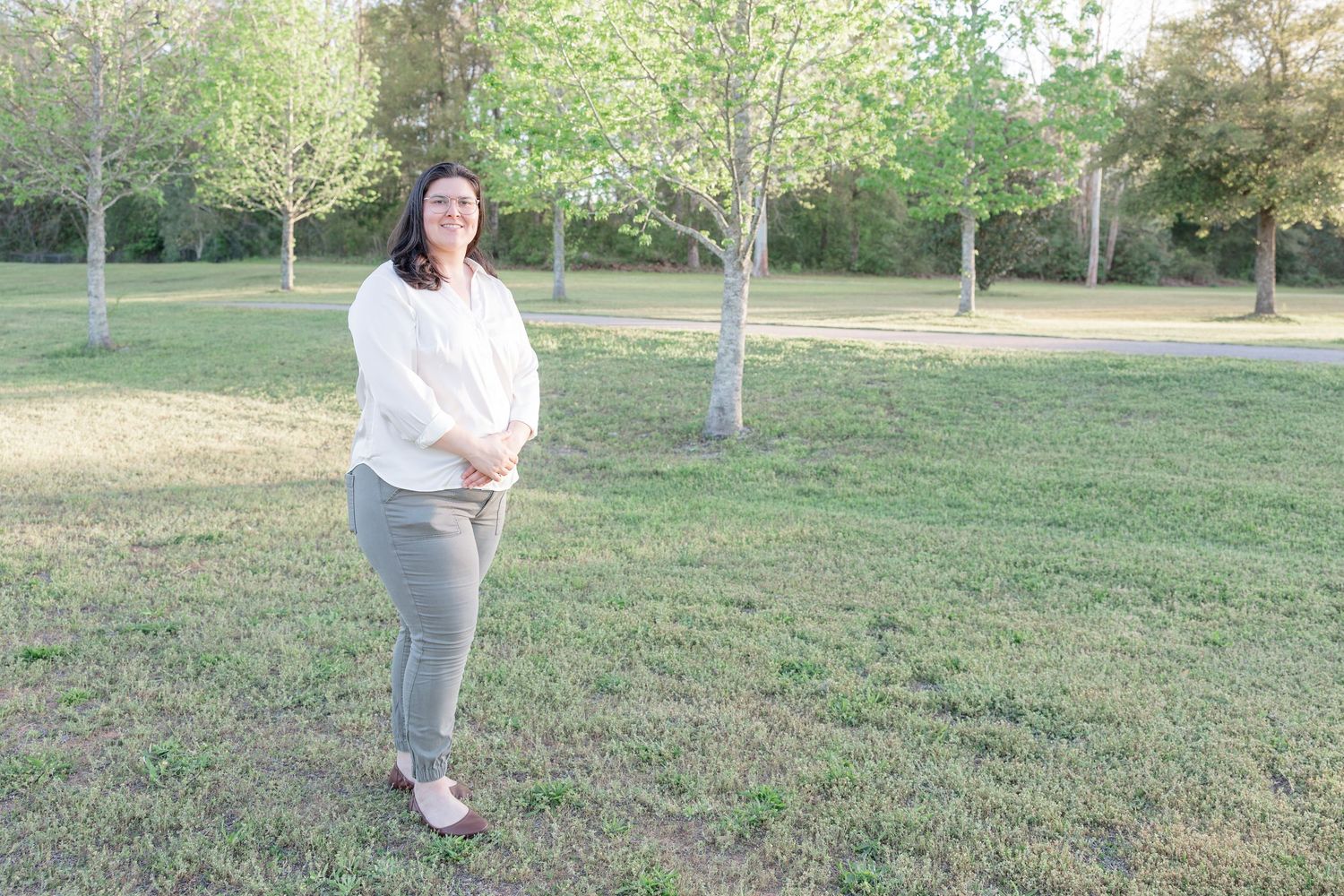 Woman in white blouse and khaki pants standing in a sunny green park with trees in the background.