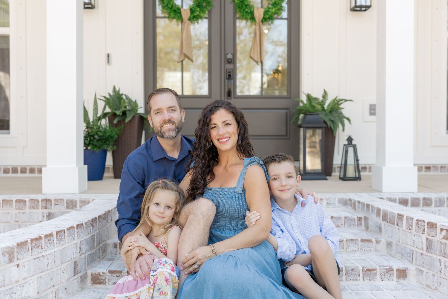 Family of four smiling on brick front porch steps of white home with dark door and greenery.