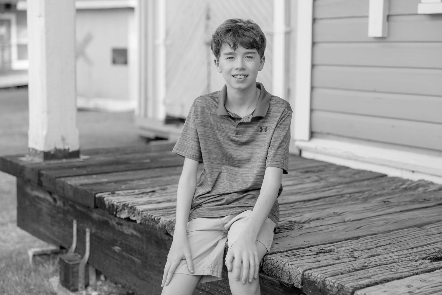 Smiling boy sitting on a rustic wooden deck outside a building in a black and white photo.