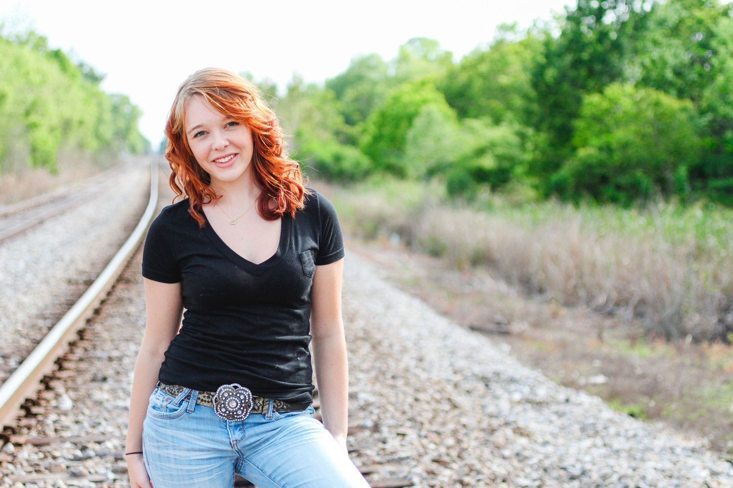 A person in a black shirt sits casually on railroad tracks surrounded by greenery and sunlight.