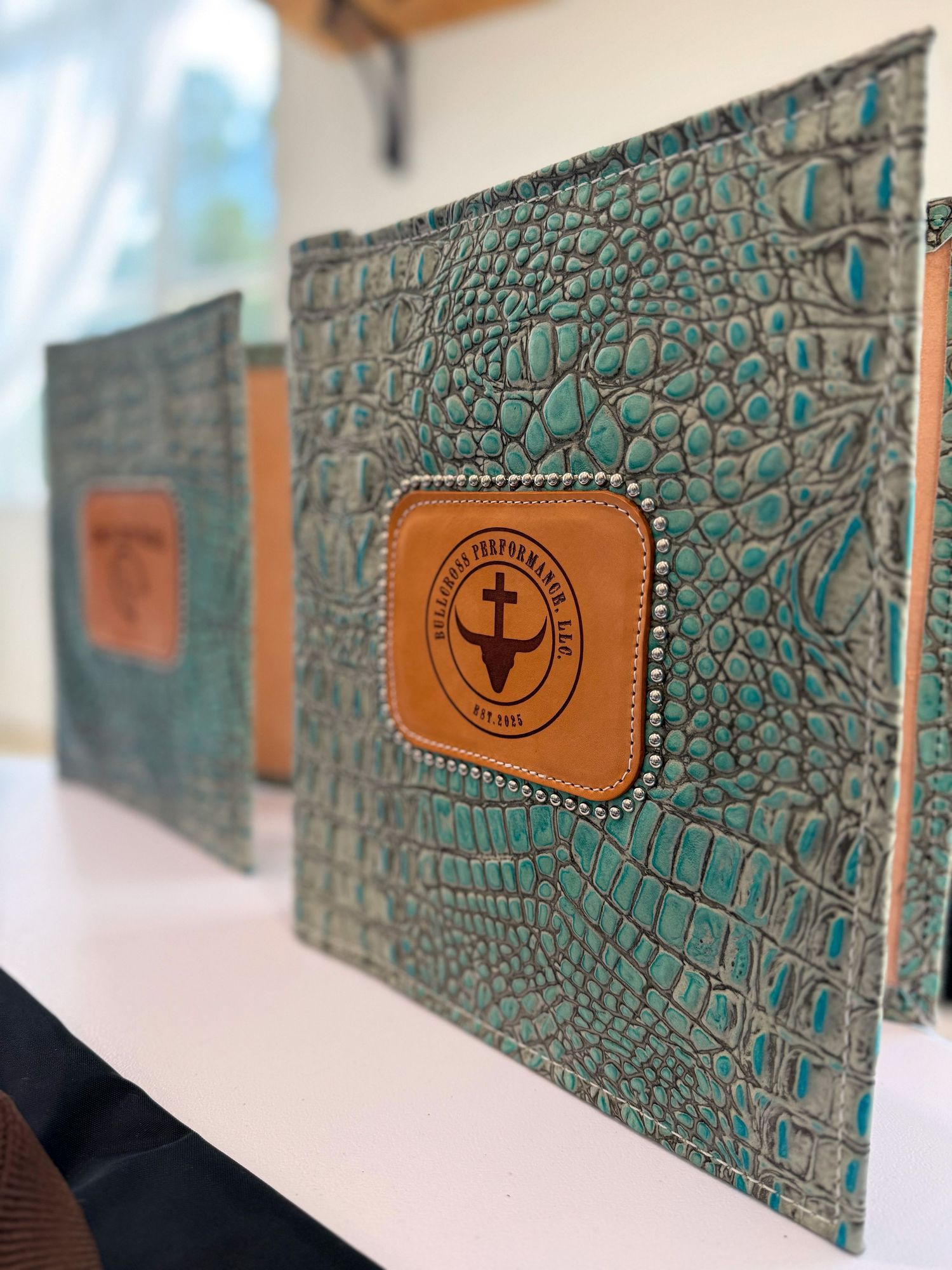Series of turquoise crocodile-textured leather wallets with tan leather accents and branded logos on white surface.
