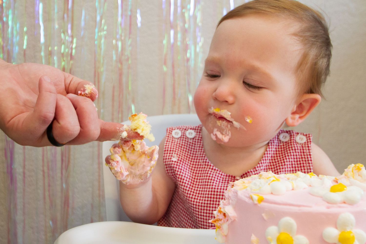 A child in a red gingham dress enjoys birthday cake while someone helps feed them at a celebration.