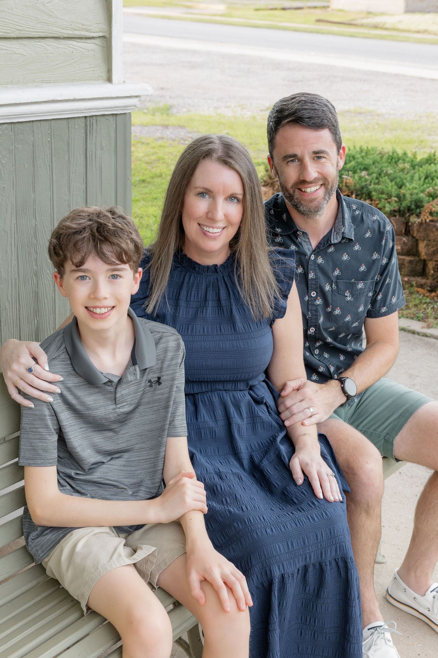 Family of three smiling on a porch, with a teenage boy, mother in blue dress, and father in patterned shirt.