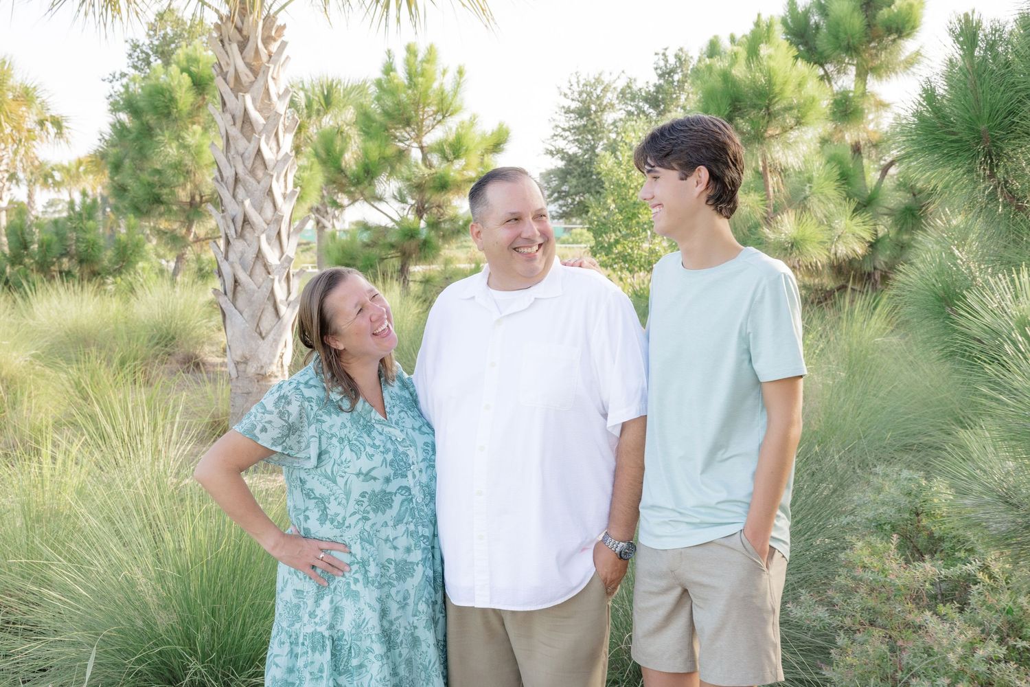 Family of three laughing together outdoors surrounded by palm trees and green shrubs.