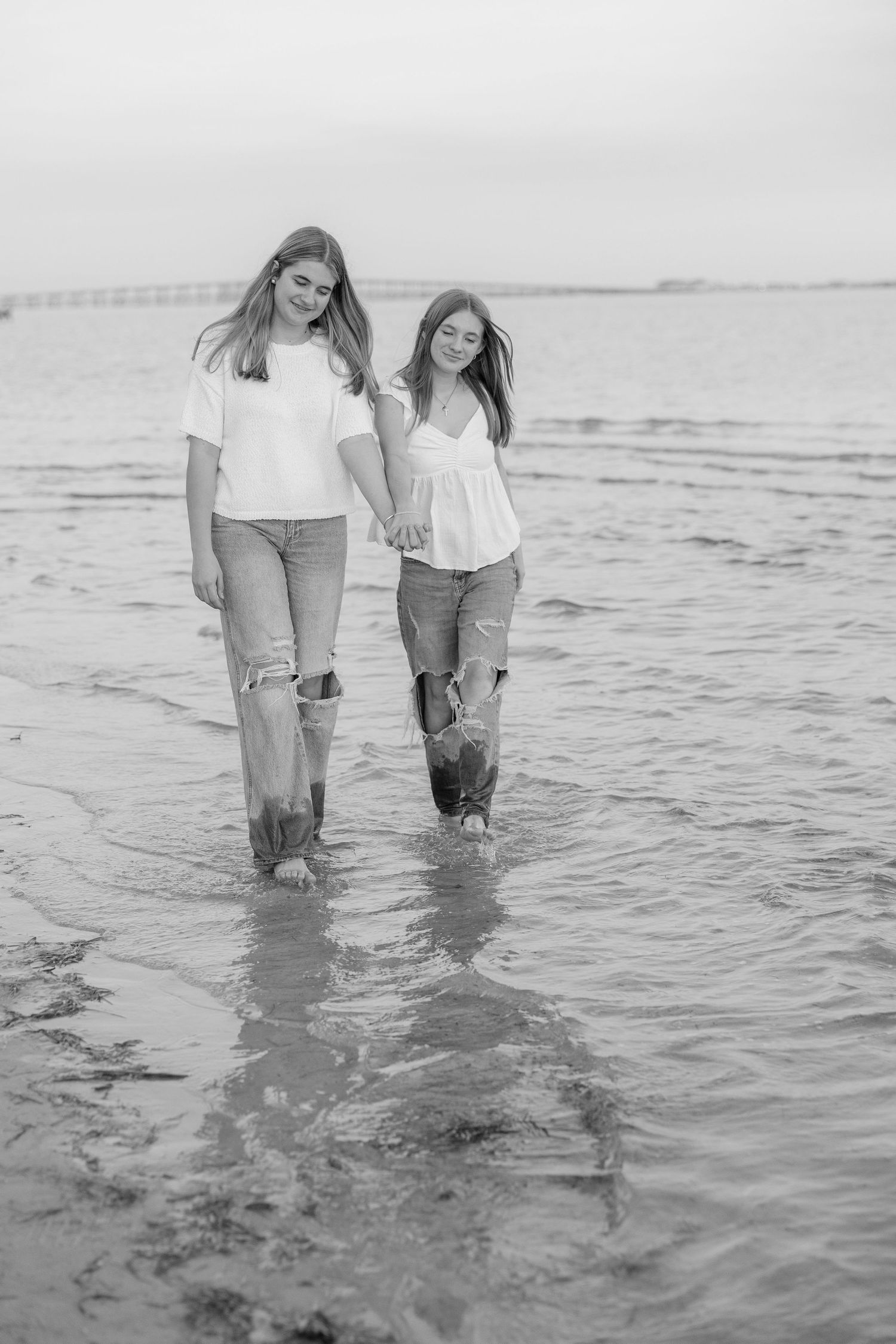 Two people walking together in shallow ocean water at the beach in a black and white photograph.
