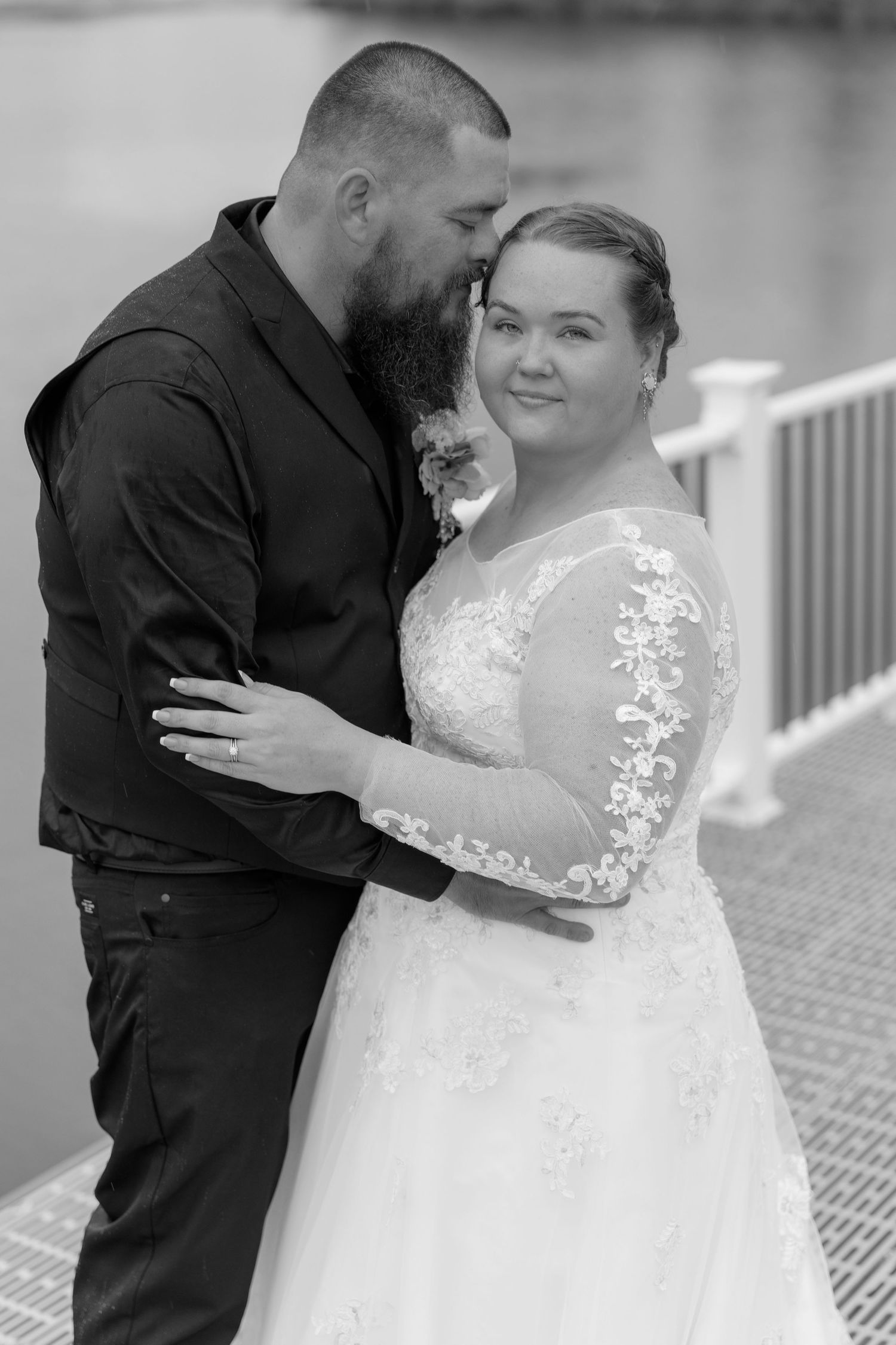 Black and white wedding photo of bearded groom in black shirt embracing bride in lace gown near waterfront railing.