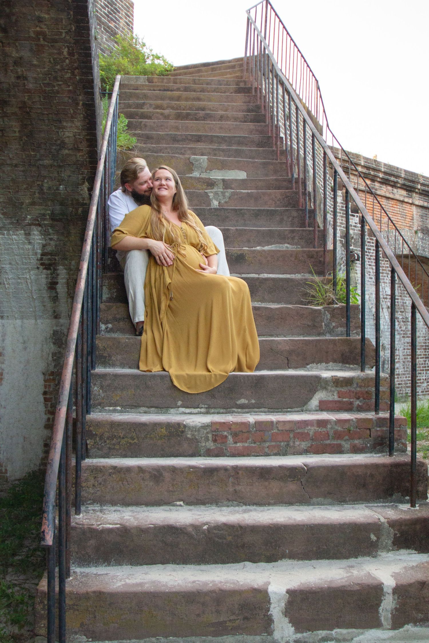 A couple sits together on outdoor stone steps with brick walls and metal railings in an urban setting.