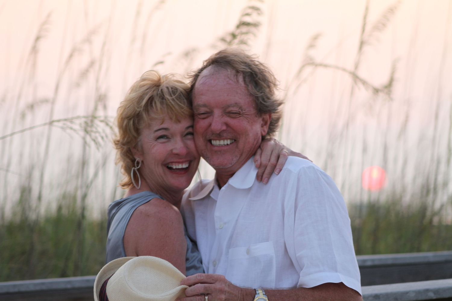 A loving couple embraces and laughs together on a beach boardwalk with sea grass in the background at sunset.