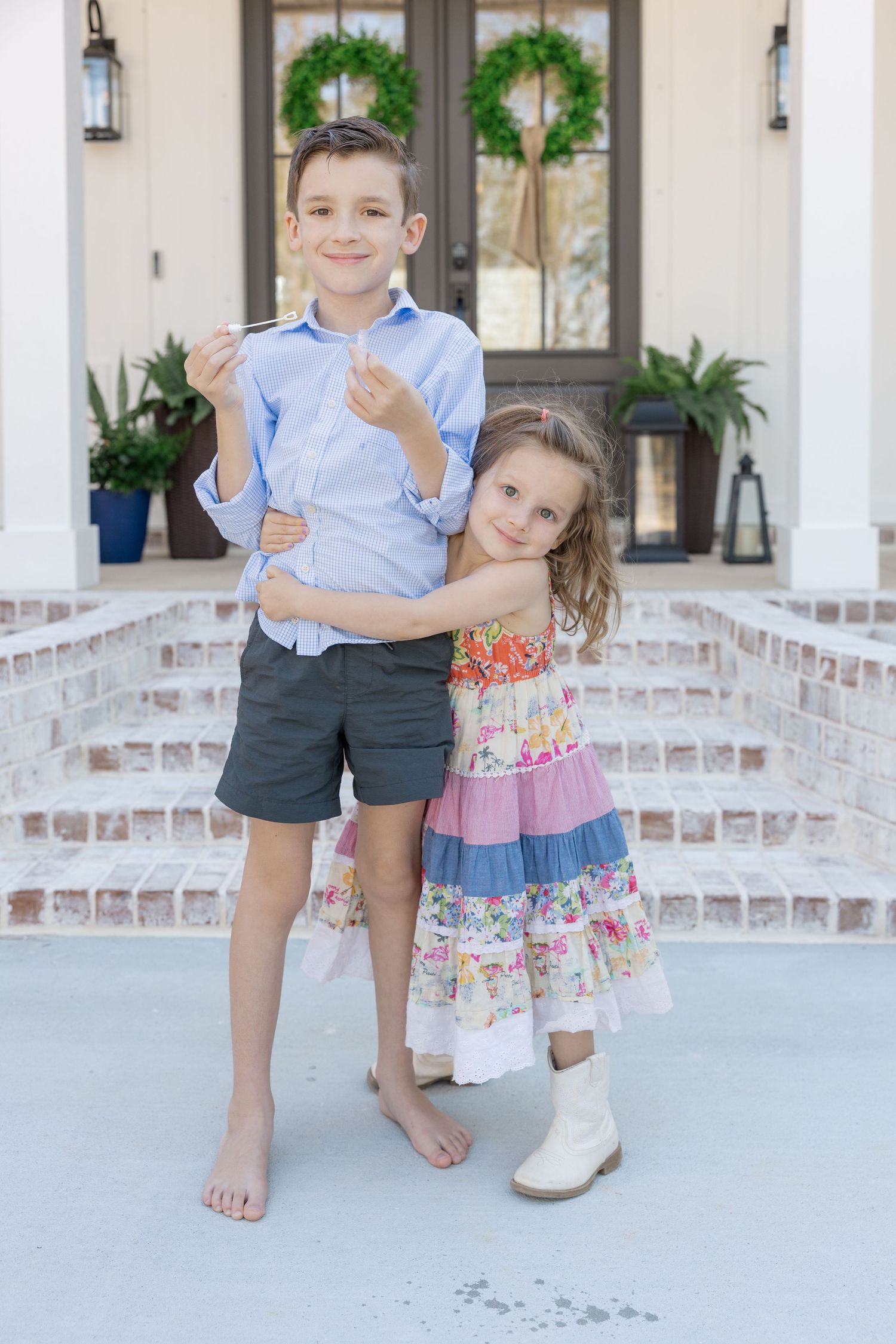Two children pose on brick steps outside a home with wreaths, girl in floral dress hugging boy in blue shirt.