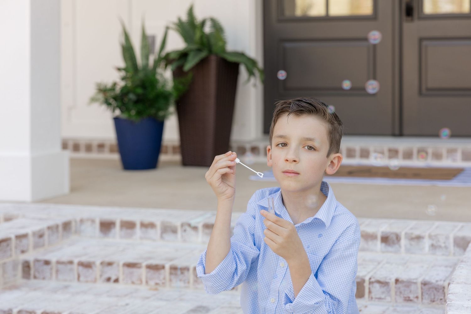 A young boy in a blue shirt blowing bubbles on a front porch steps outdoors.