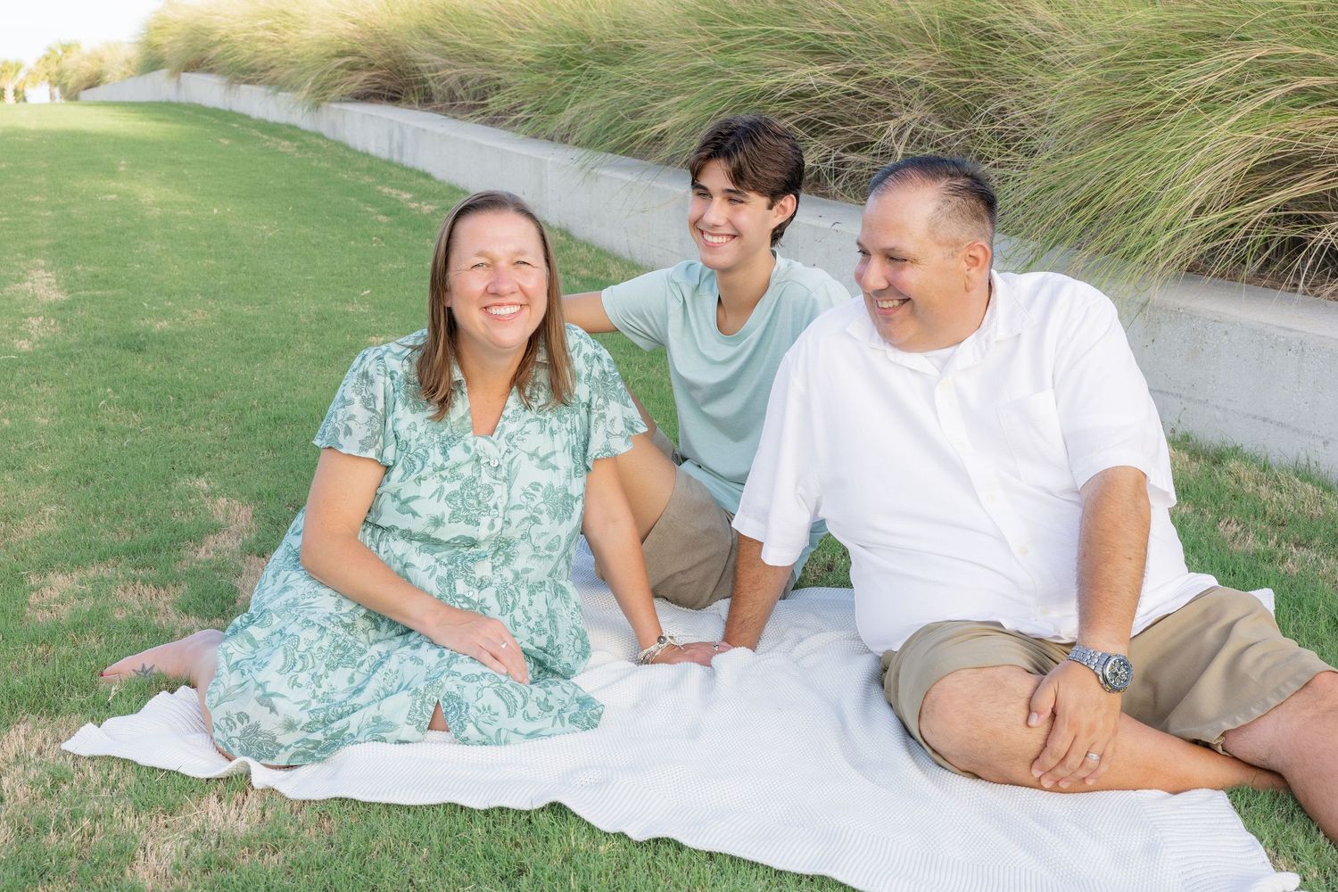 Family of three laughing and sitting on a white blanket on green grass near tall ornamental grasses.