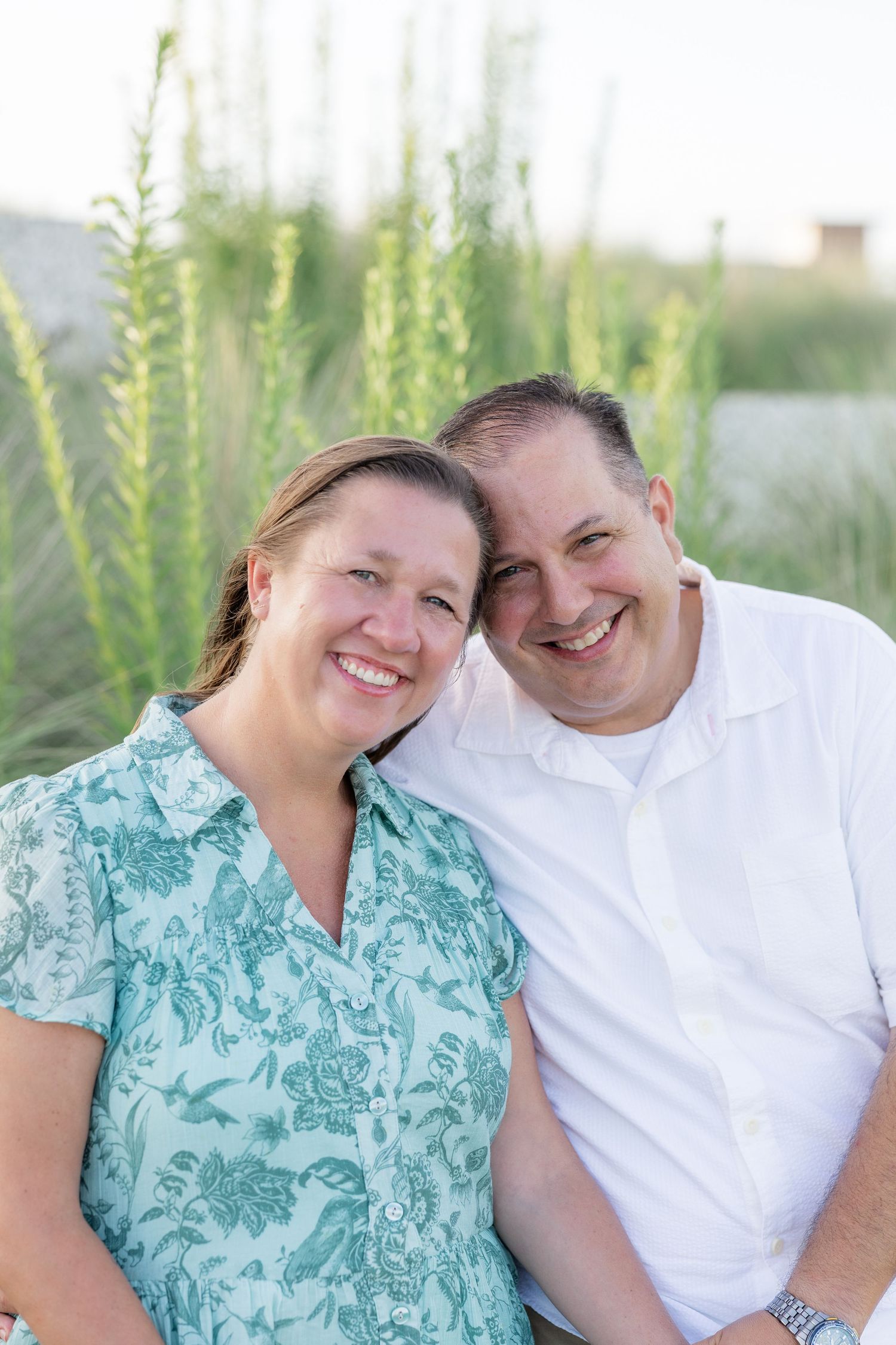 Smiling couple posing outdoors near tall green sea grass, woman in teal floral dress and man in white shirt.