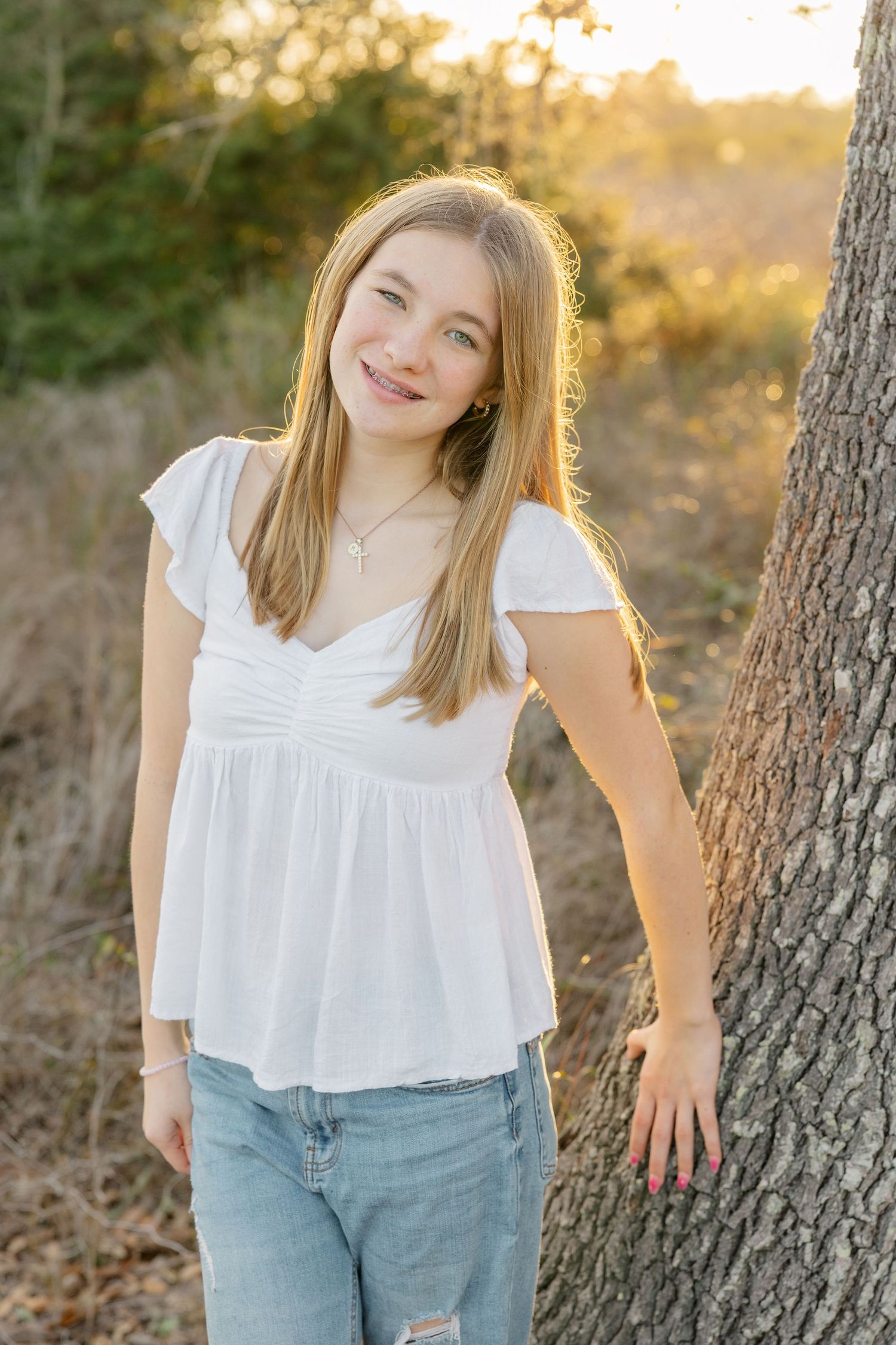 Young person in white top and jeans poses casually against trees during golden hour sunset.