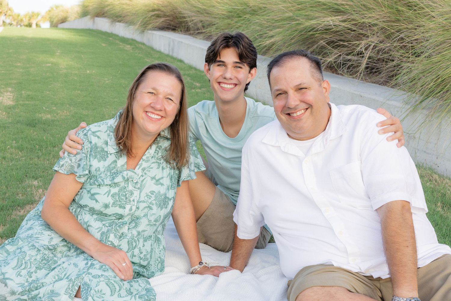 Happy family of three sitting together on green grass lawn smiling outdoors.