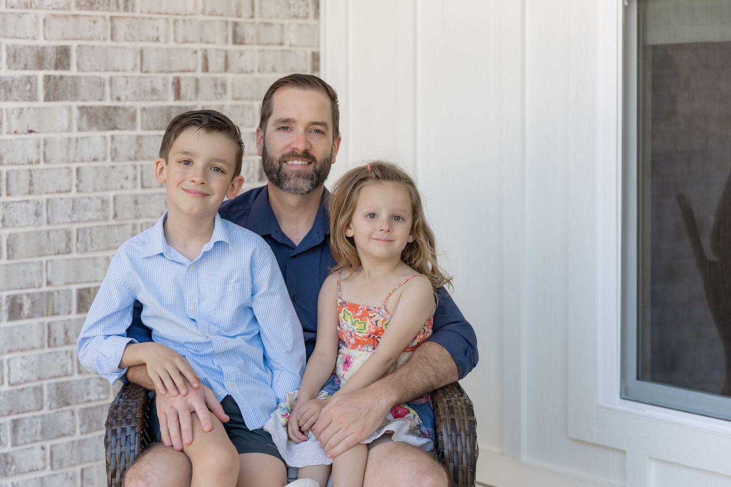 A smiling father sits with his two young children on a porch, posing for a family portrait near a brick wall.