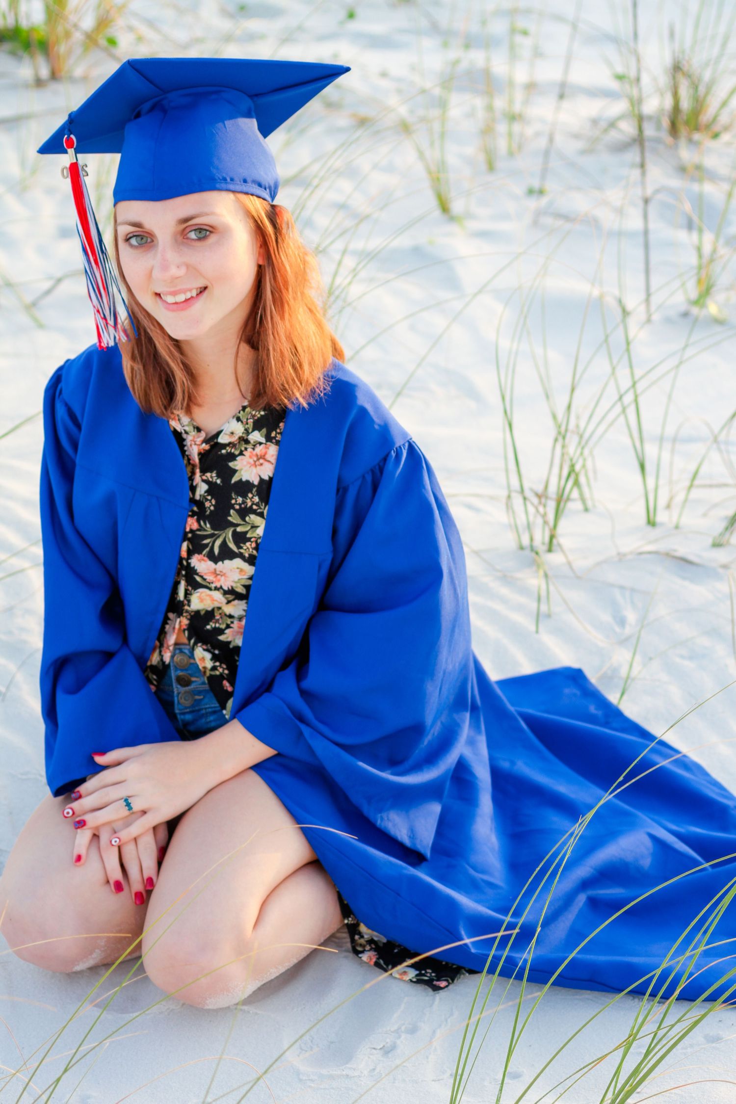 Graduate in bright blue cap and gown poses on white sandy beach with sea grass on sunny day.