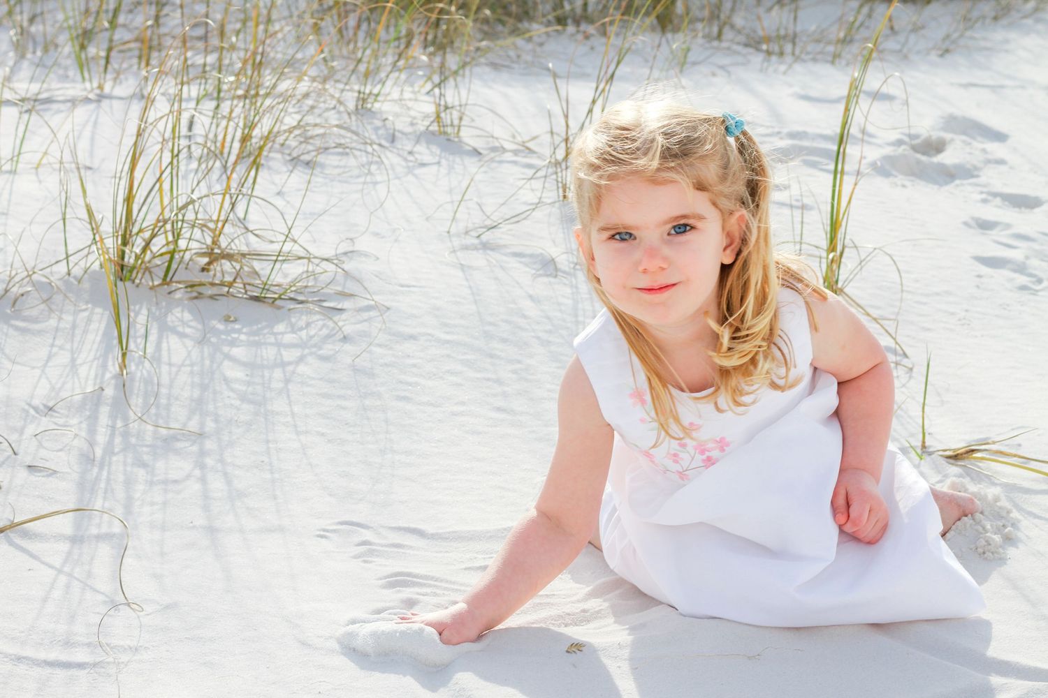 A child in a white dress sits among the sand dunes and sea grass at a sunny beach.
