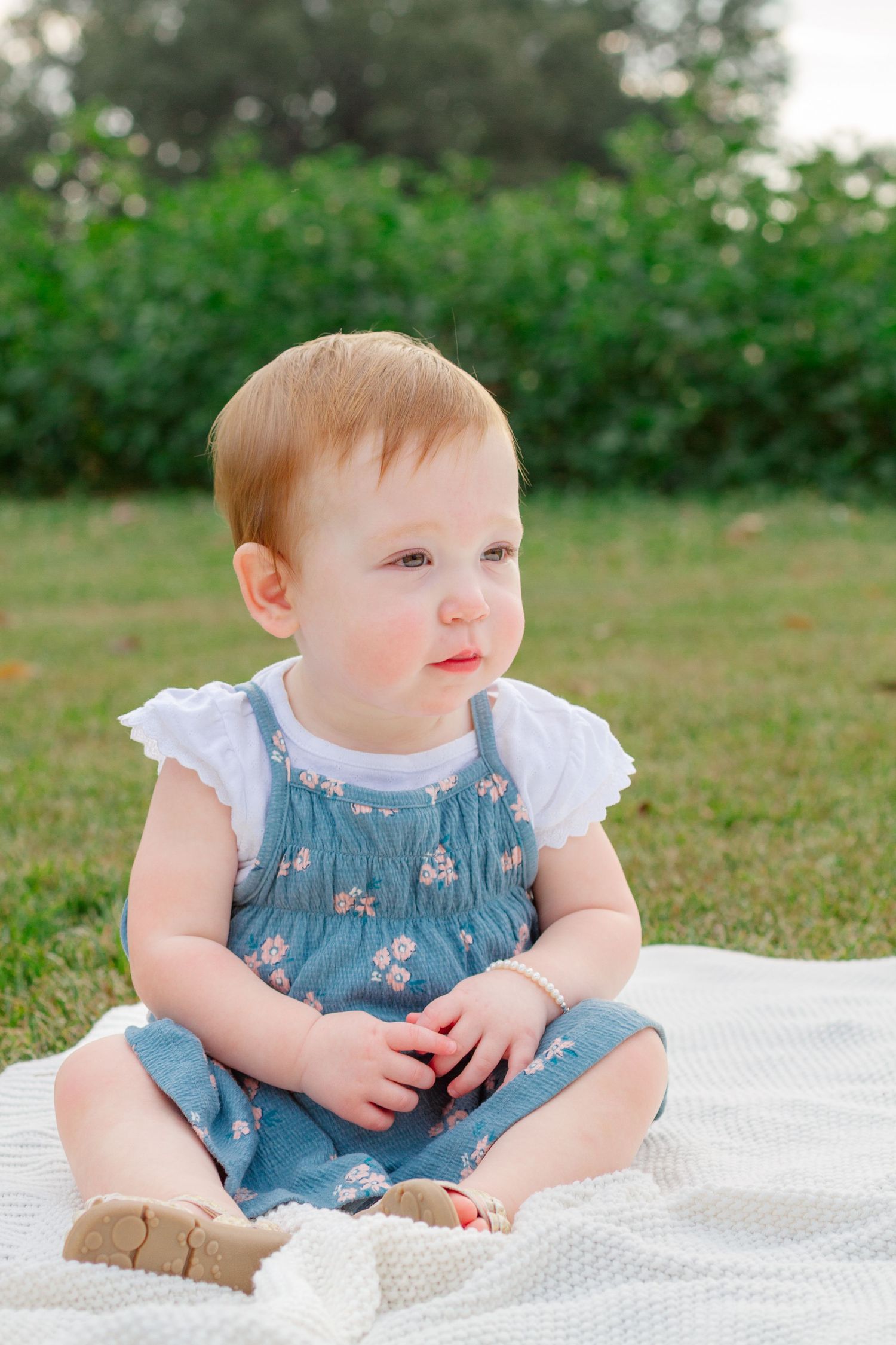 Baby in floral denim overalls sits on white blanket outdoors during summer photo session.