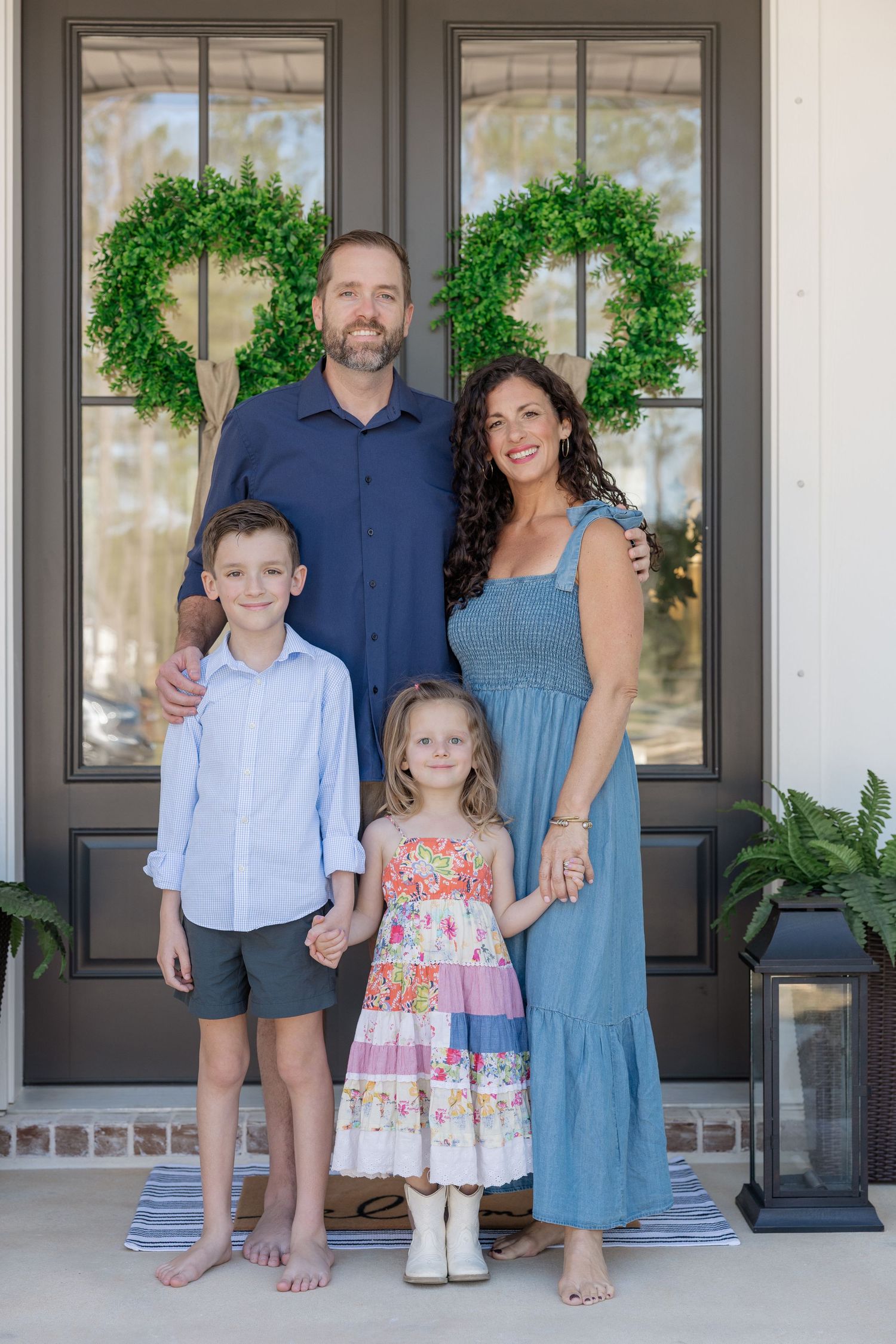 Family of four posing in front of dark double doors with green wreaths on a white home's front porch.