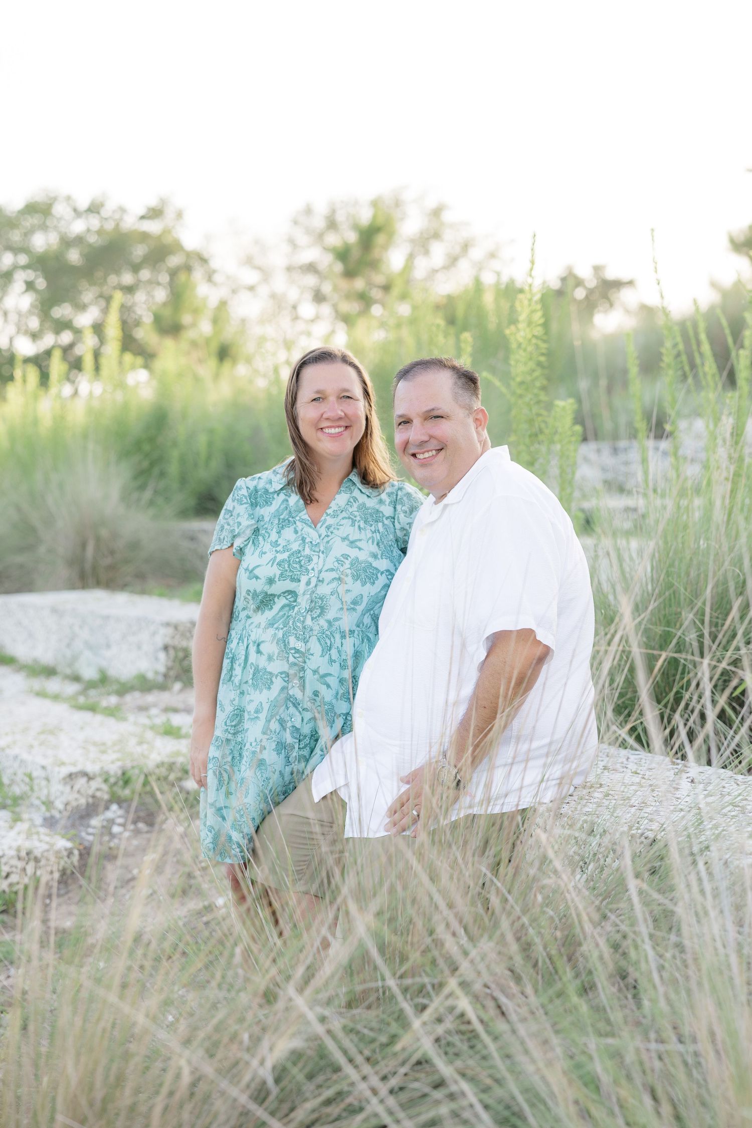 Smiling couple standing outdoors among tall green grasses, woman in teal dress and man in white shirt.
