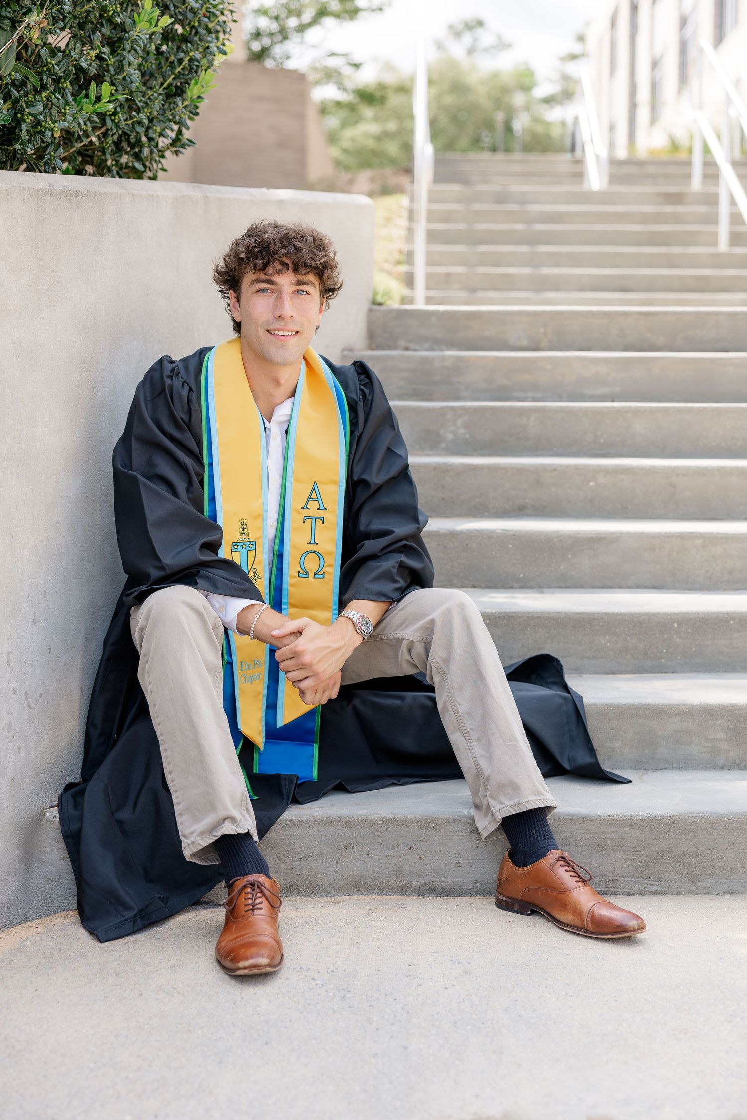 Young man in graduation gown and Alpha Tau Omega stole sits on concrete steps smiling for senior photos.