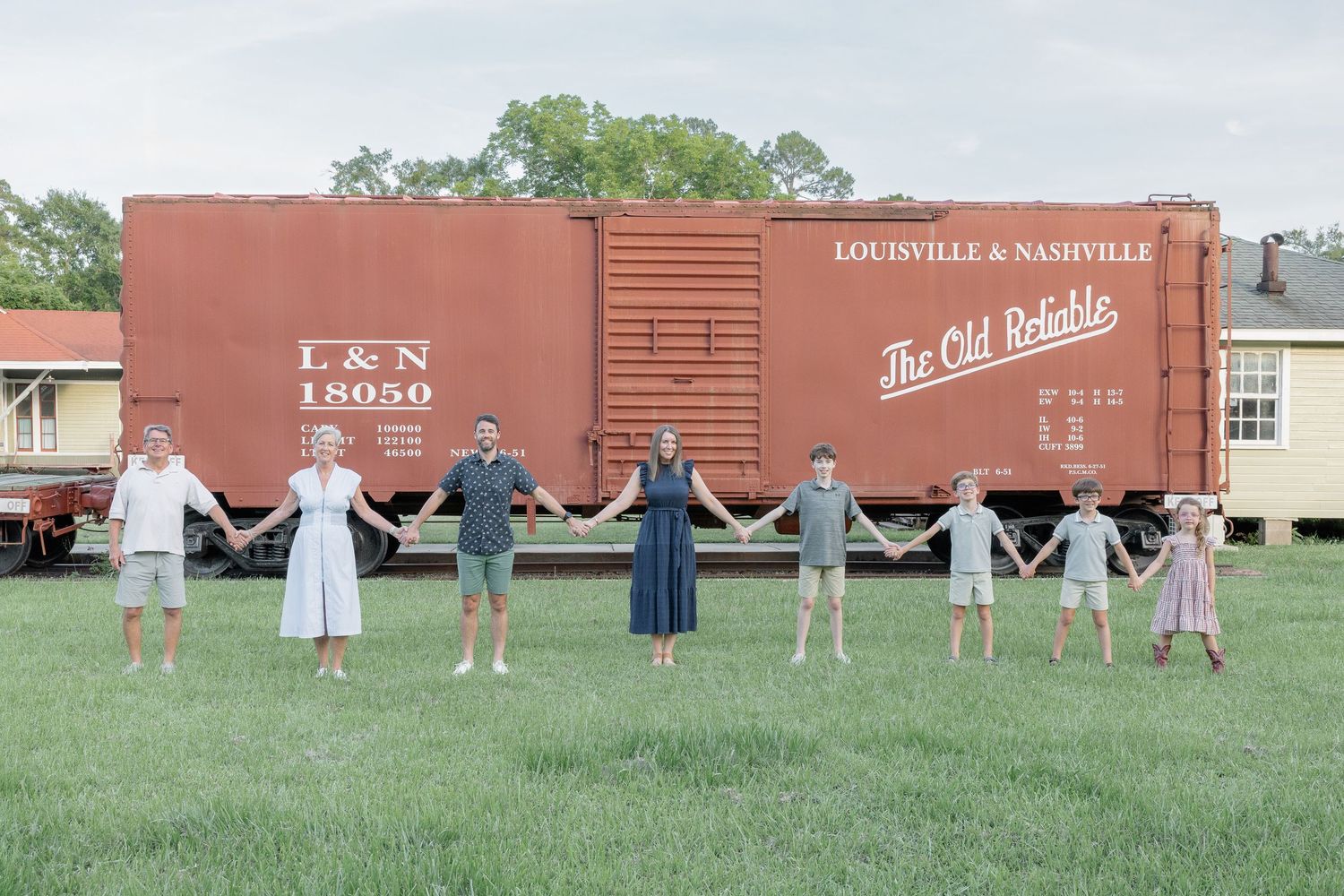 Family of eight holding hands in a line in front of a vintage Louisville & Nashville railroad boxcar on green grass.