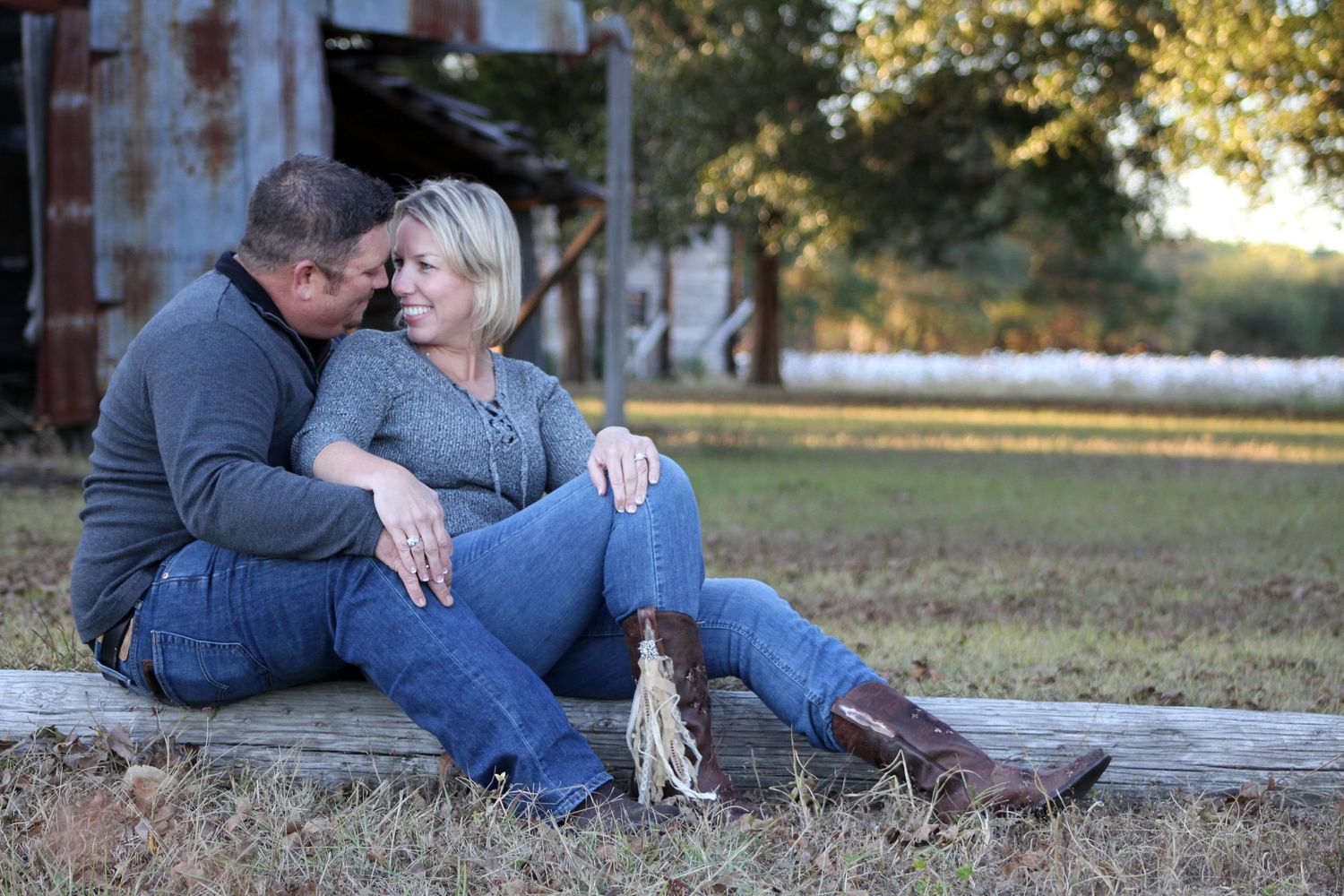 A couple in blue jeans and casual wear sits intimately on a rustic log in an outdoor setting during golden hour.