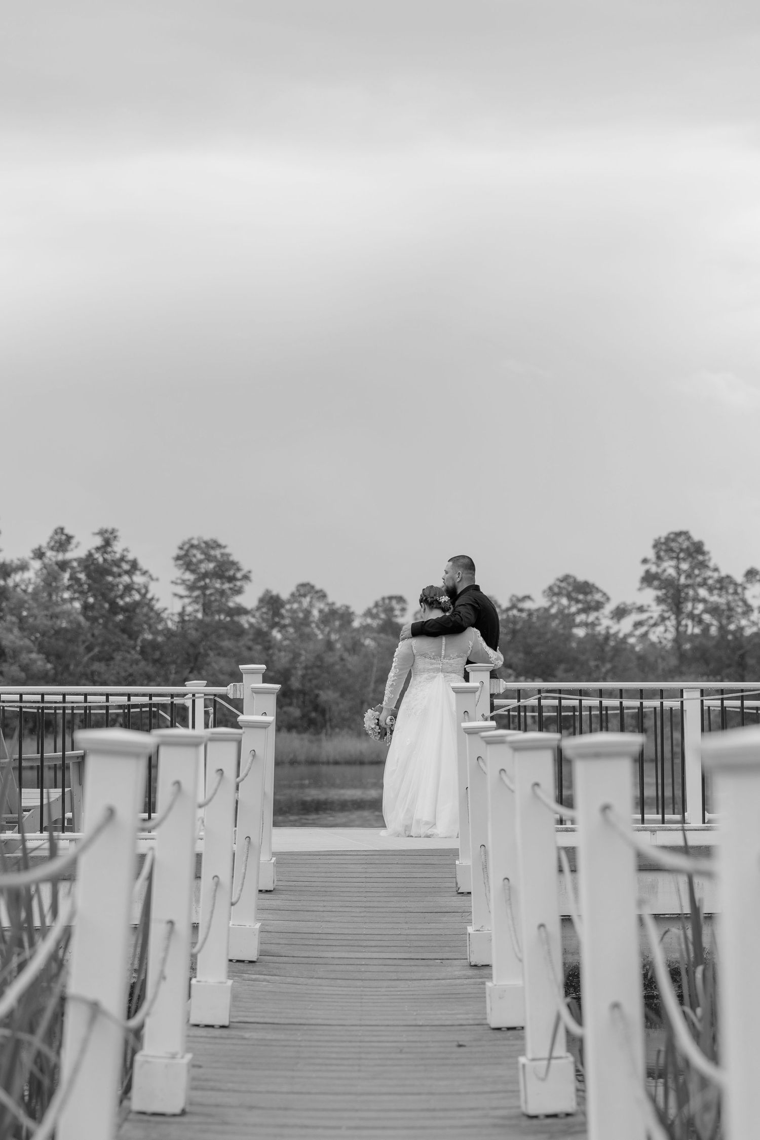Black and white photo of a bride and groom embracing on a wooden dock overlooking a marsh landscape.