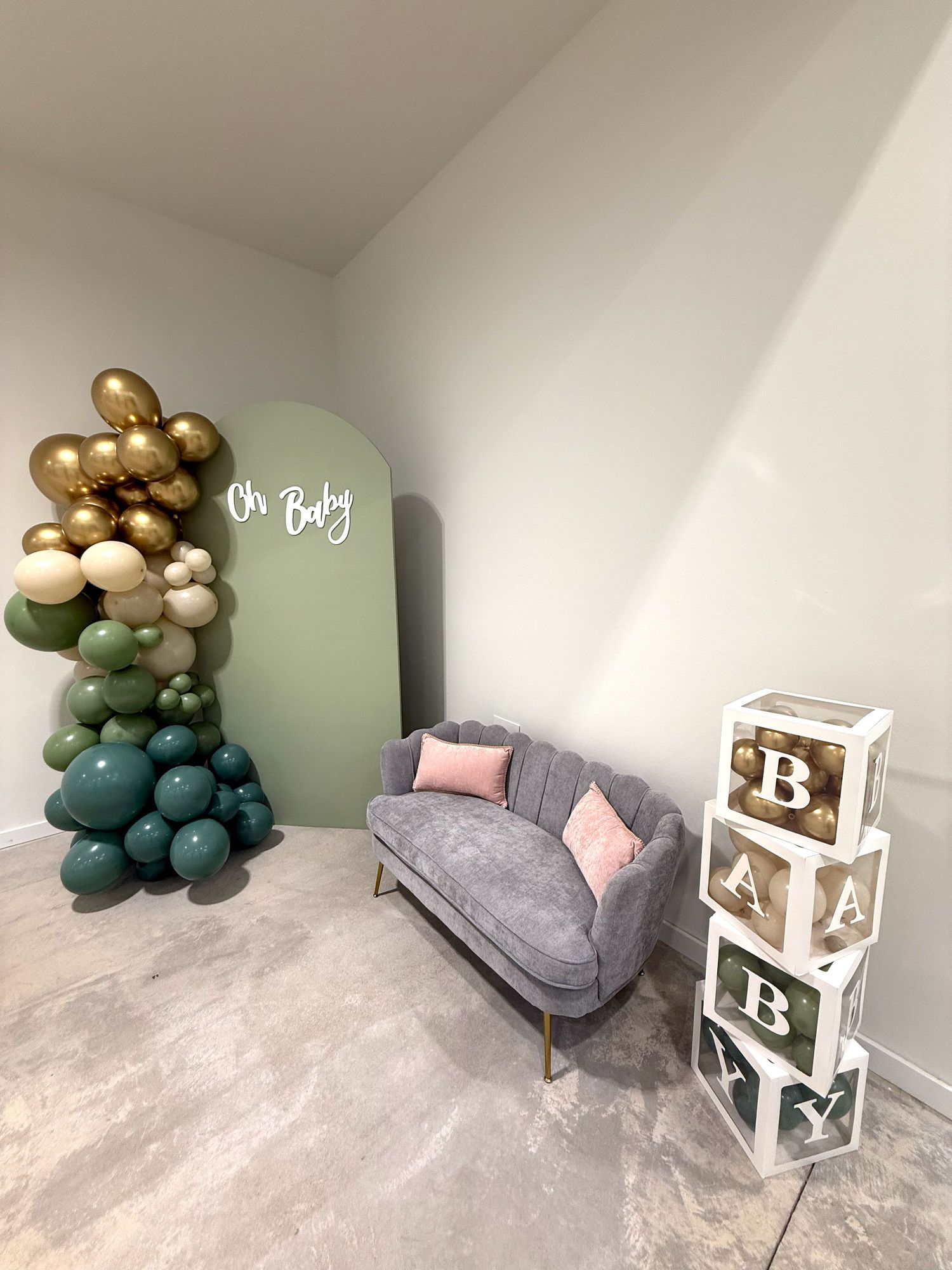 Modern baby shower setup featuring a gray armchair, decorative balloon arch in green and gold, and white alphabet blocks.