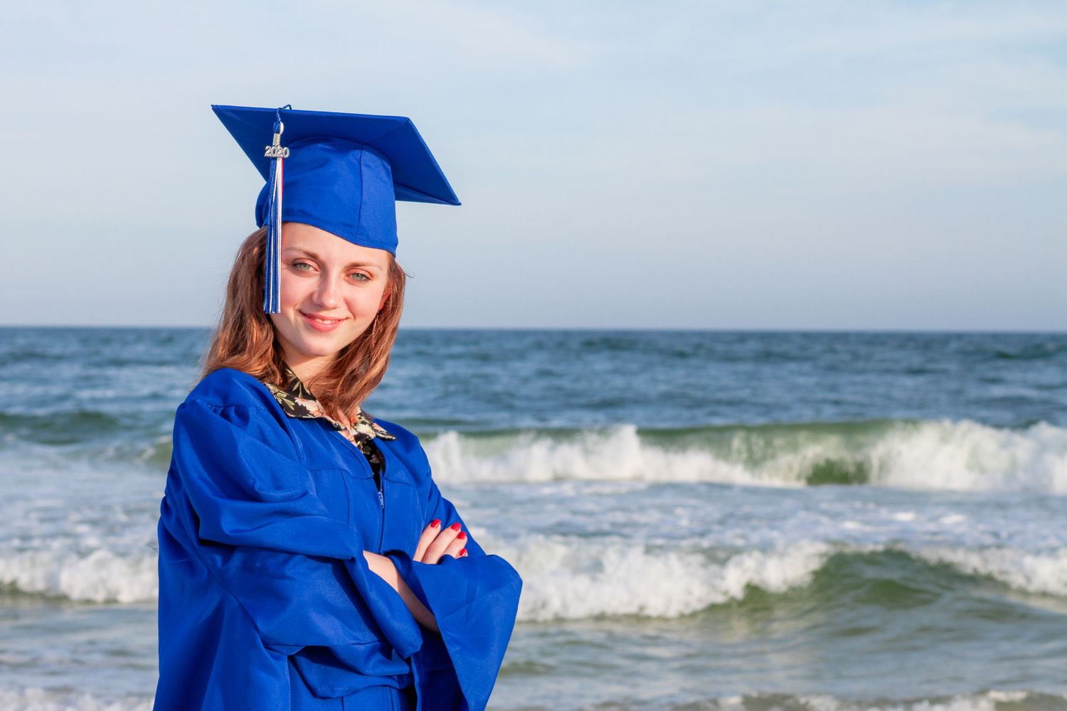 Graduate wearing blue cap and gown posing confidently with arms crossed on sunny beach with ocean waves in background.