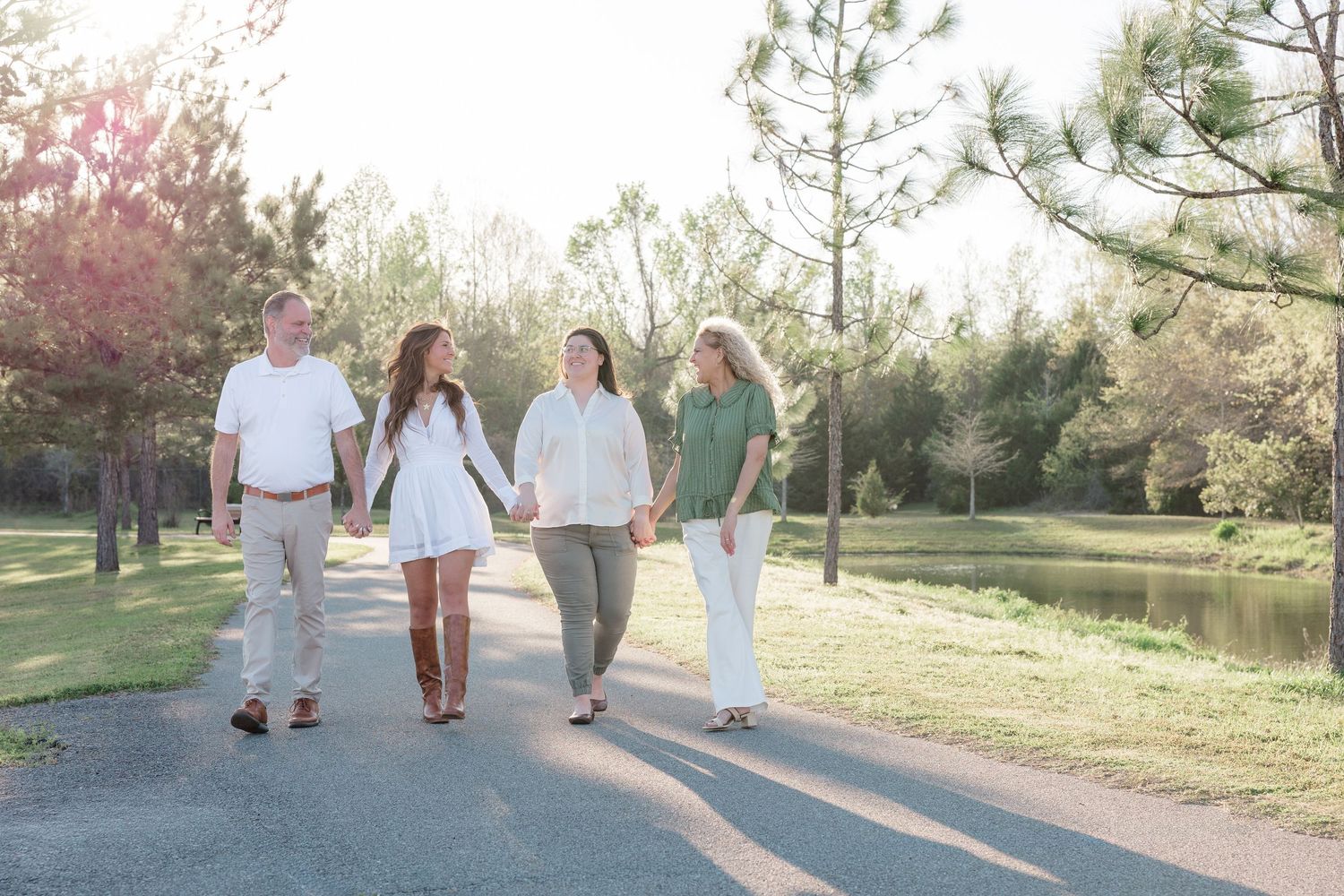 Family of four holding hands walking on a sunlit park path surrounded by trees and a pond.