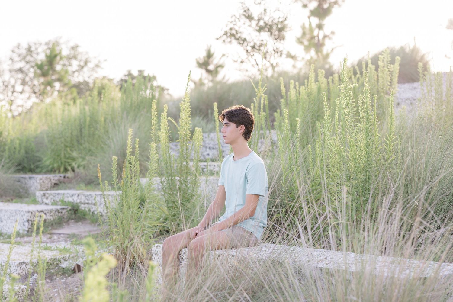 A young man in a mint green shirt sits peacefully on a rock surrounded by tall wild grasses in a serene outdoor setting.