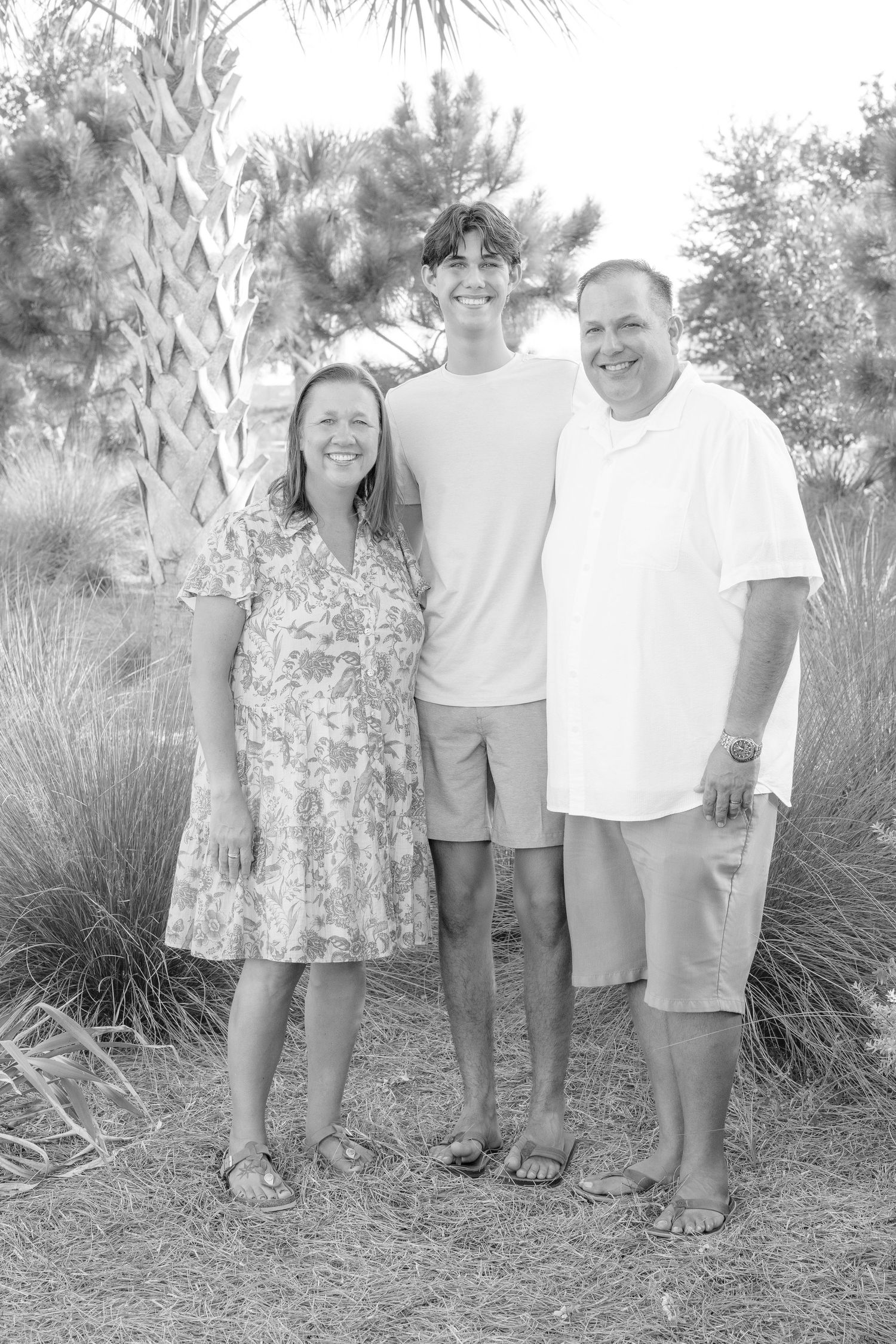 Black and white family portrait of two adults and a teen standing outdoors near palm trees.