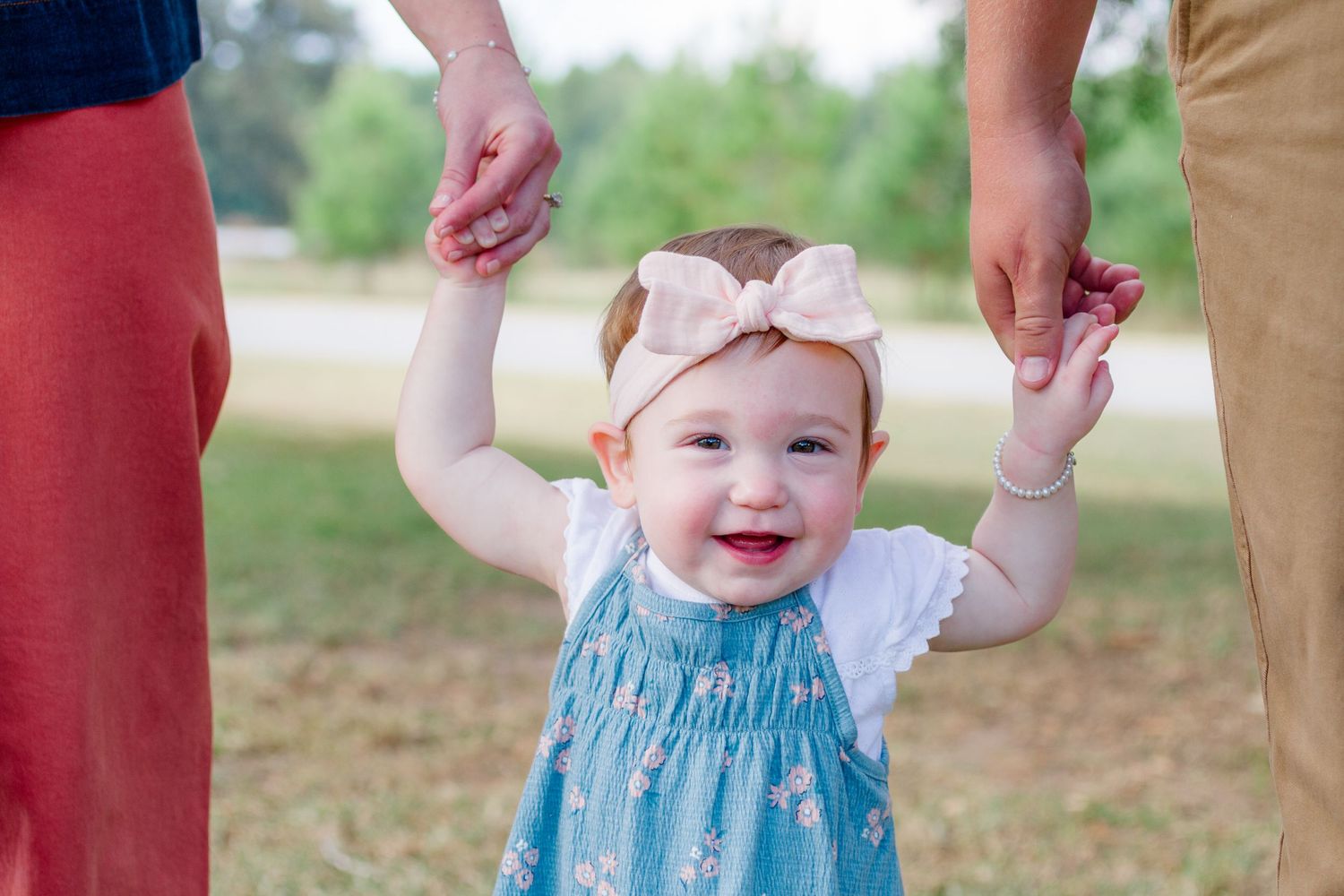 A small child wearing a blue dress smiles while being held up by adult hands in an outdoor setting.