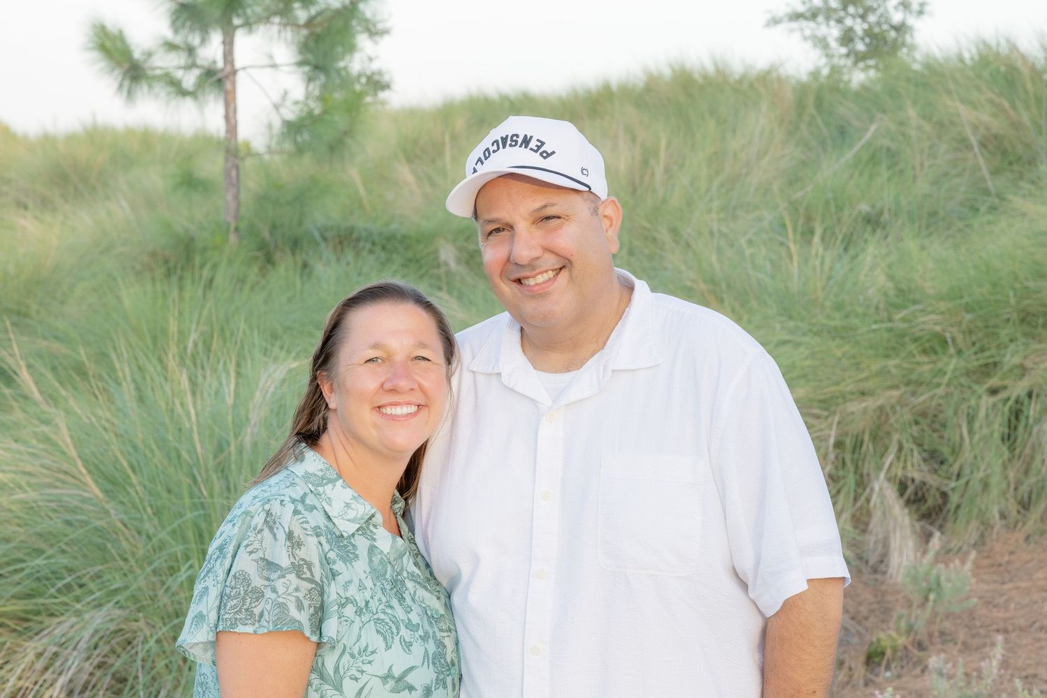 Smiling couple posing outdoors near sandy dunes with green grass, man in white shirt and cap, woman in floral top.