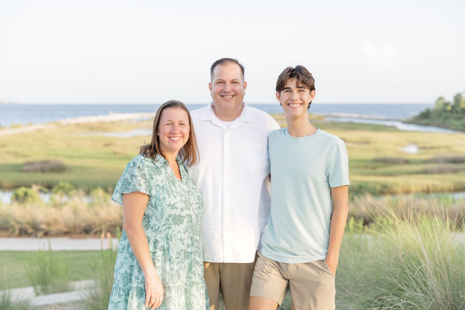 Family of three smiling together outdoors with a scenic coastal waterway and marsh in the background.