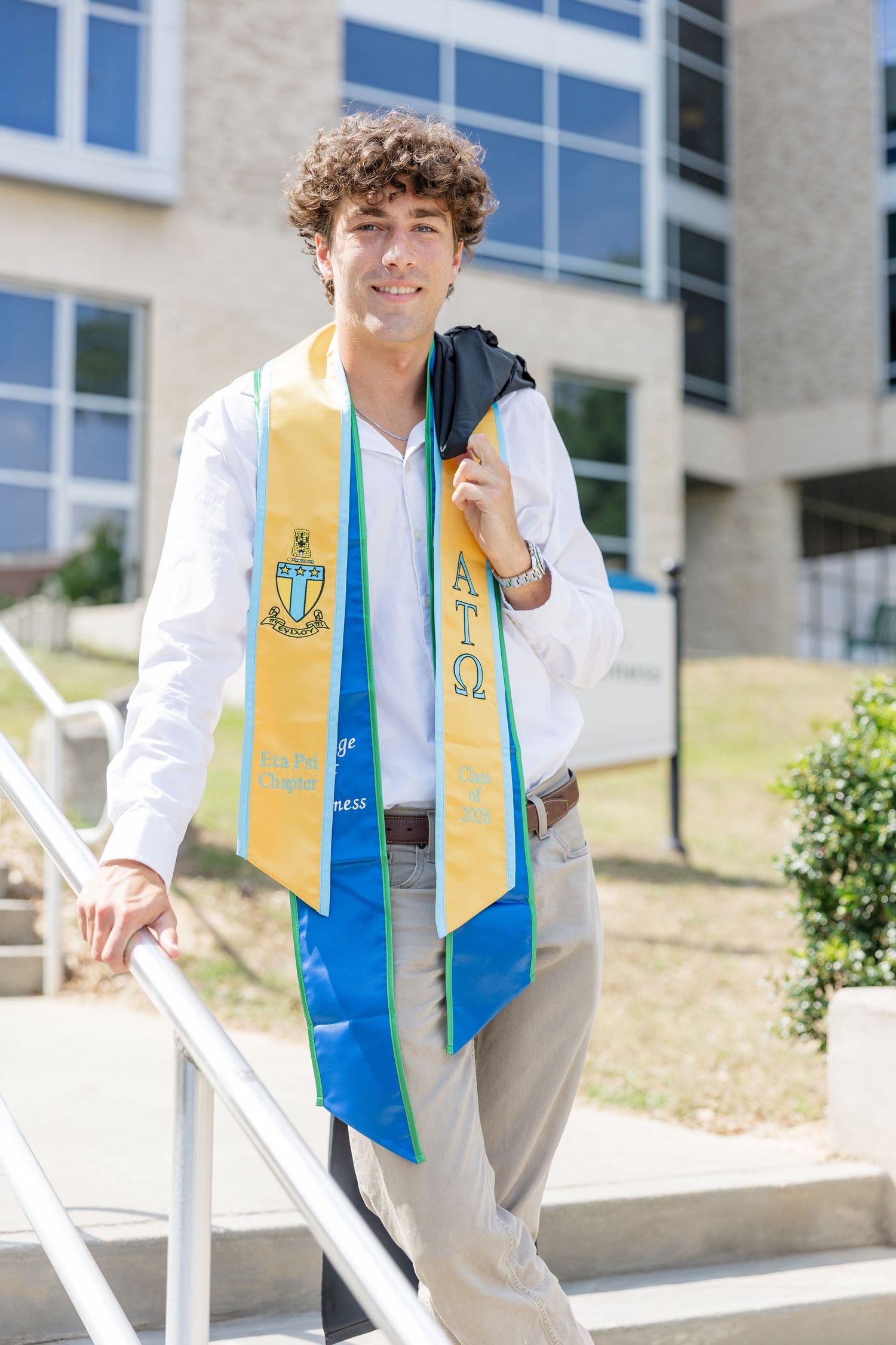 Young male college graduate smiling on steps wearing white shirt and blue and gold graduation stole with Greek letters.